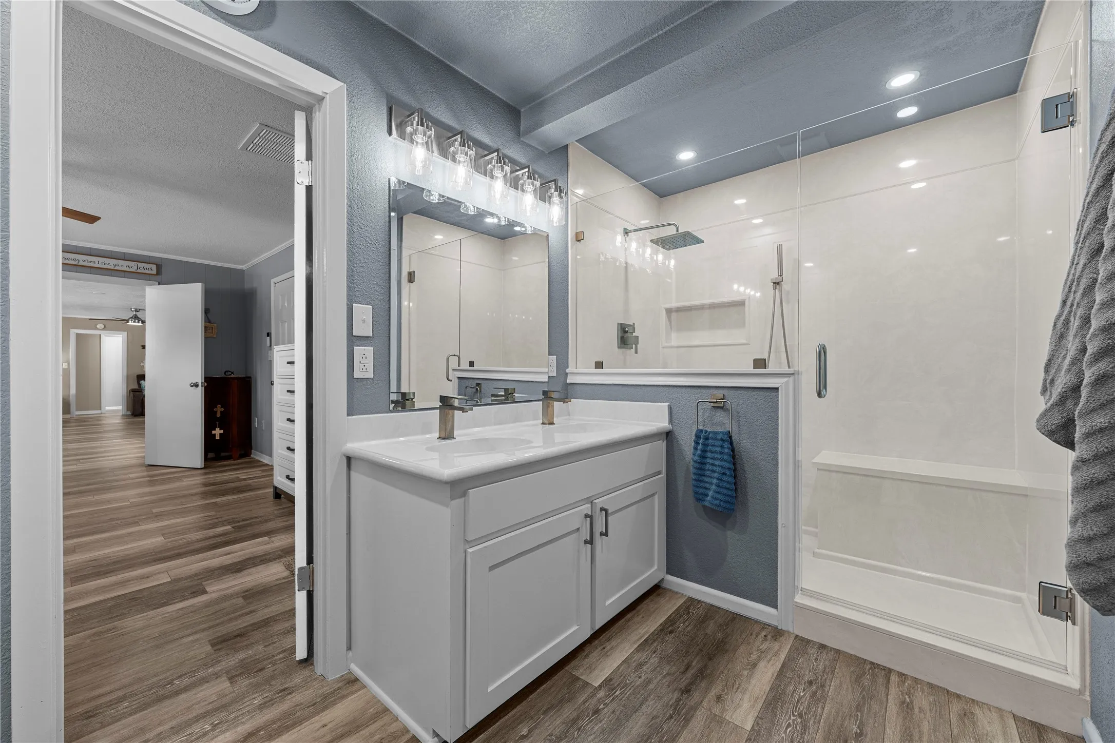Dual vanity with white cabinetry, glass-enclosed shower with rain showerhead, recessed lighting, and wood-finish flooring