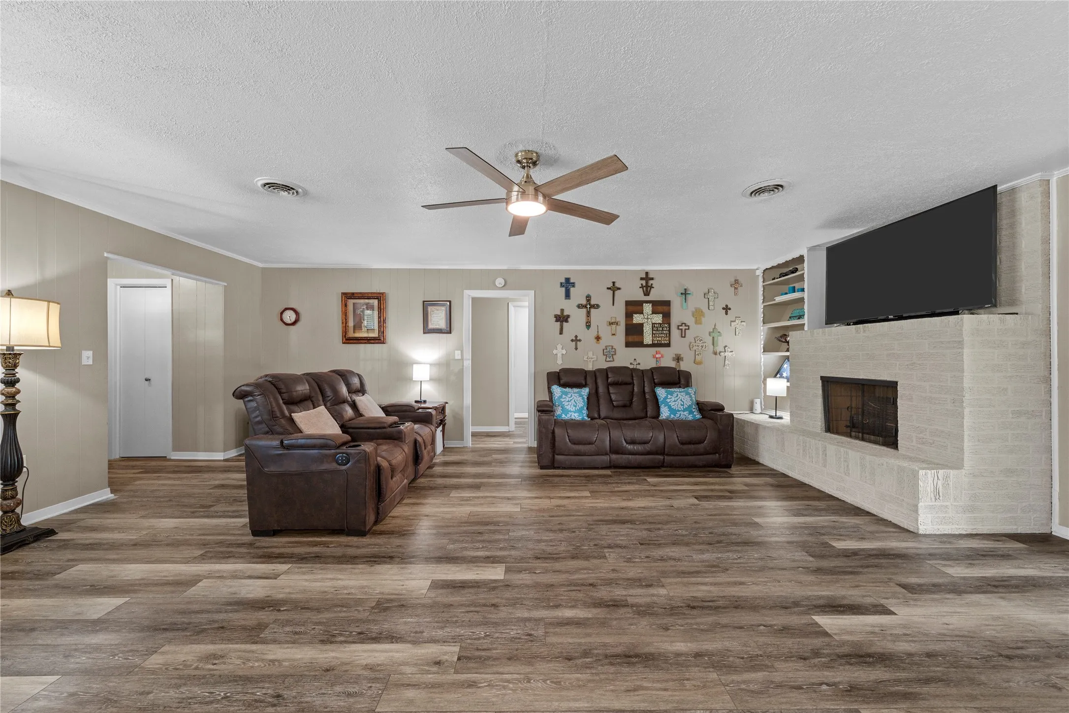 Spacious living area featuring wood-finish flooring, a white brick fireplace with a built-in hearth, recessed shelving, and a ceiling fan