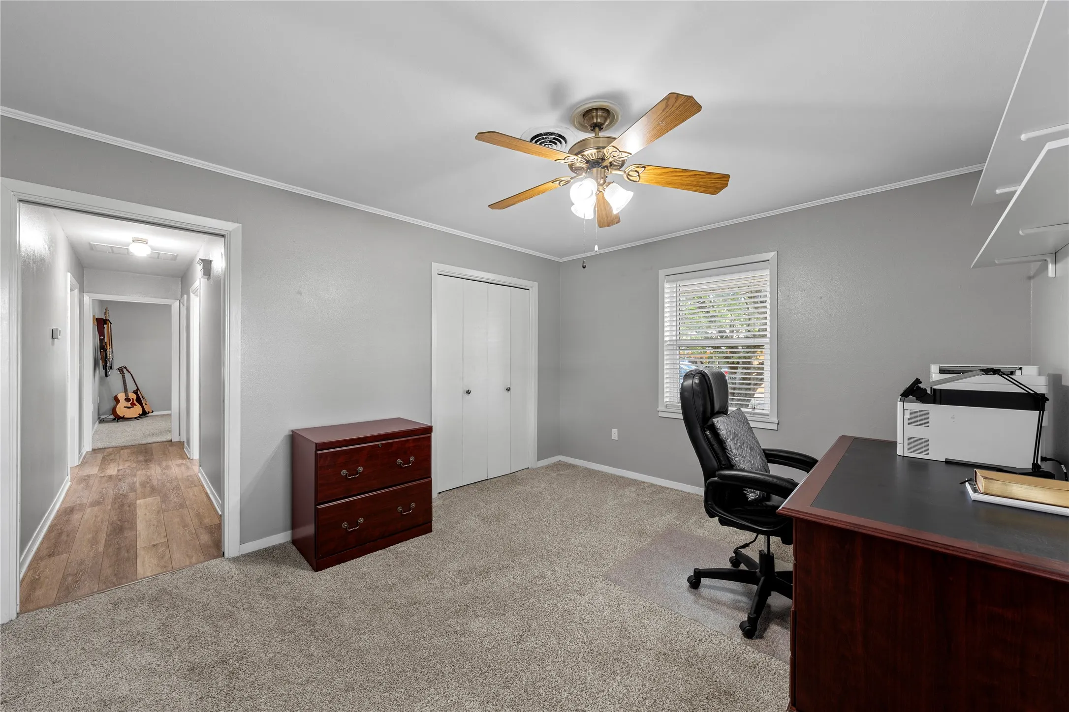 Neutral gray wall paint, textured carpet flooring, ceiling fan with integrated lighting, window with blinds, and white bi-fold closet doors