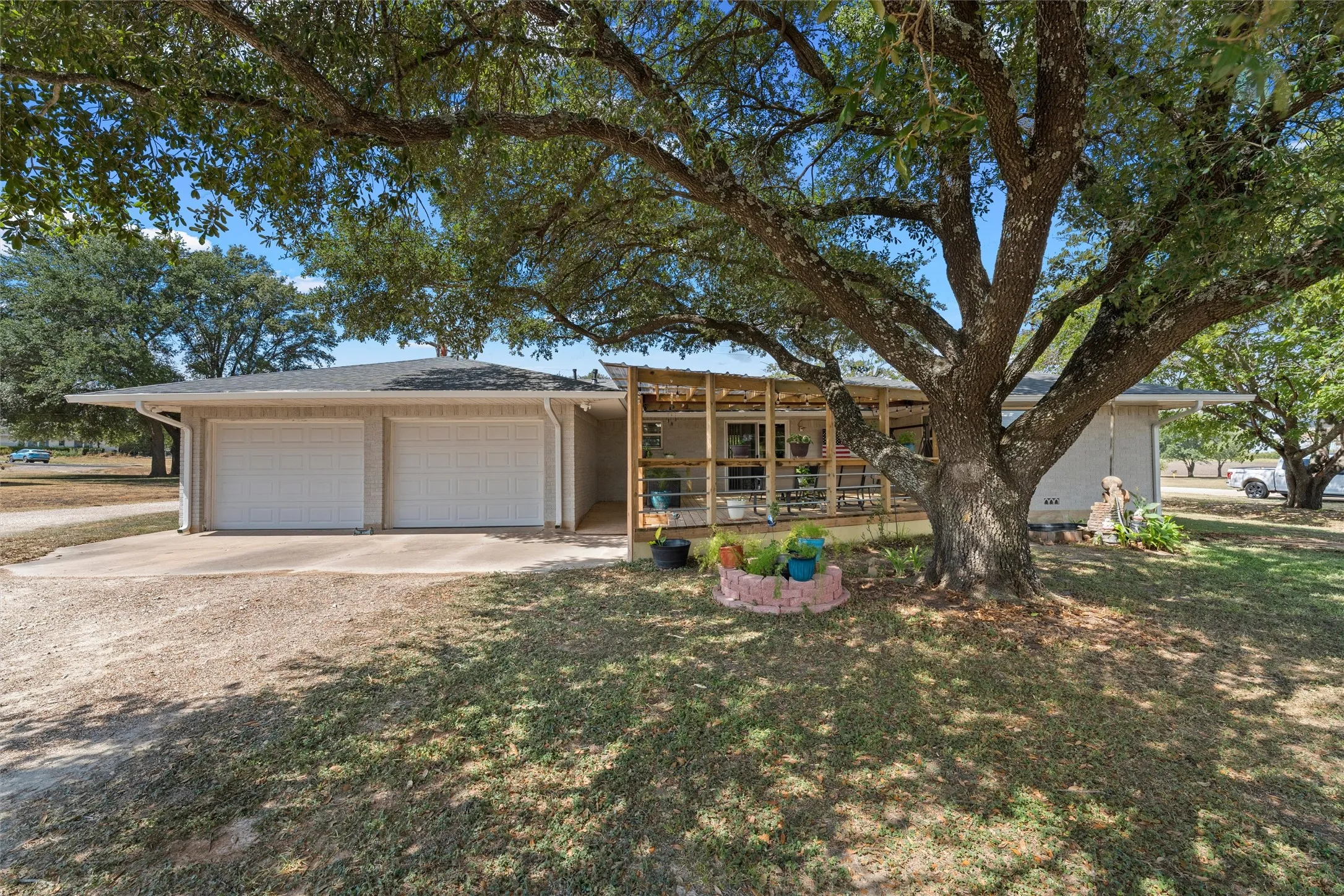 Two-car garage with white doors, expansive front yard, mature tree providing ample shade, and an extended front porch