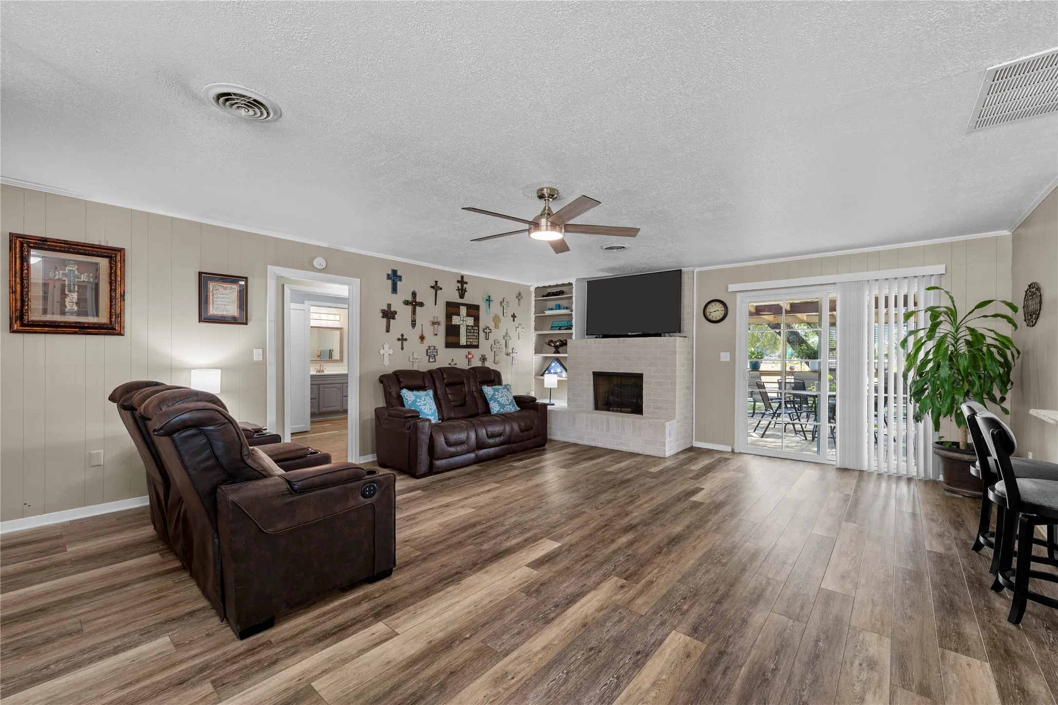 Spacious living area featuring wood-finish flooring, a white painted brick fireplace, built-in shelving, and a ceiling fan