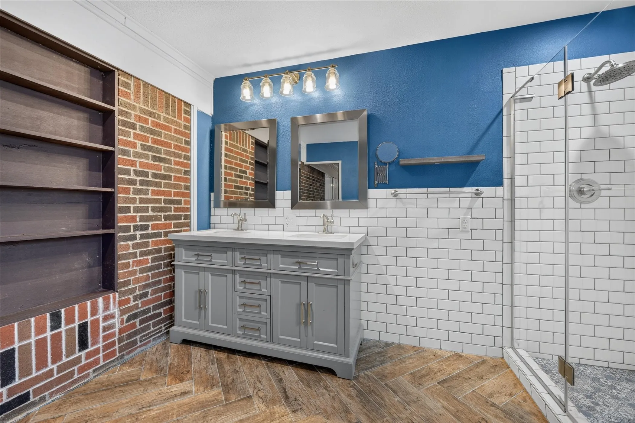 Dual-sink vanity with a light-toned countertop, herringbone wood-finish flooring, subway tile shower enclosure with clear glass, built-in shelving, and exposed brick wall detailing