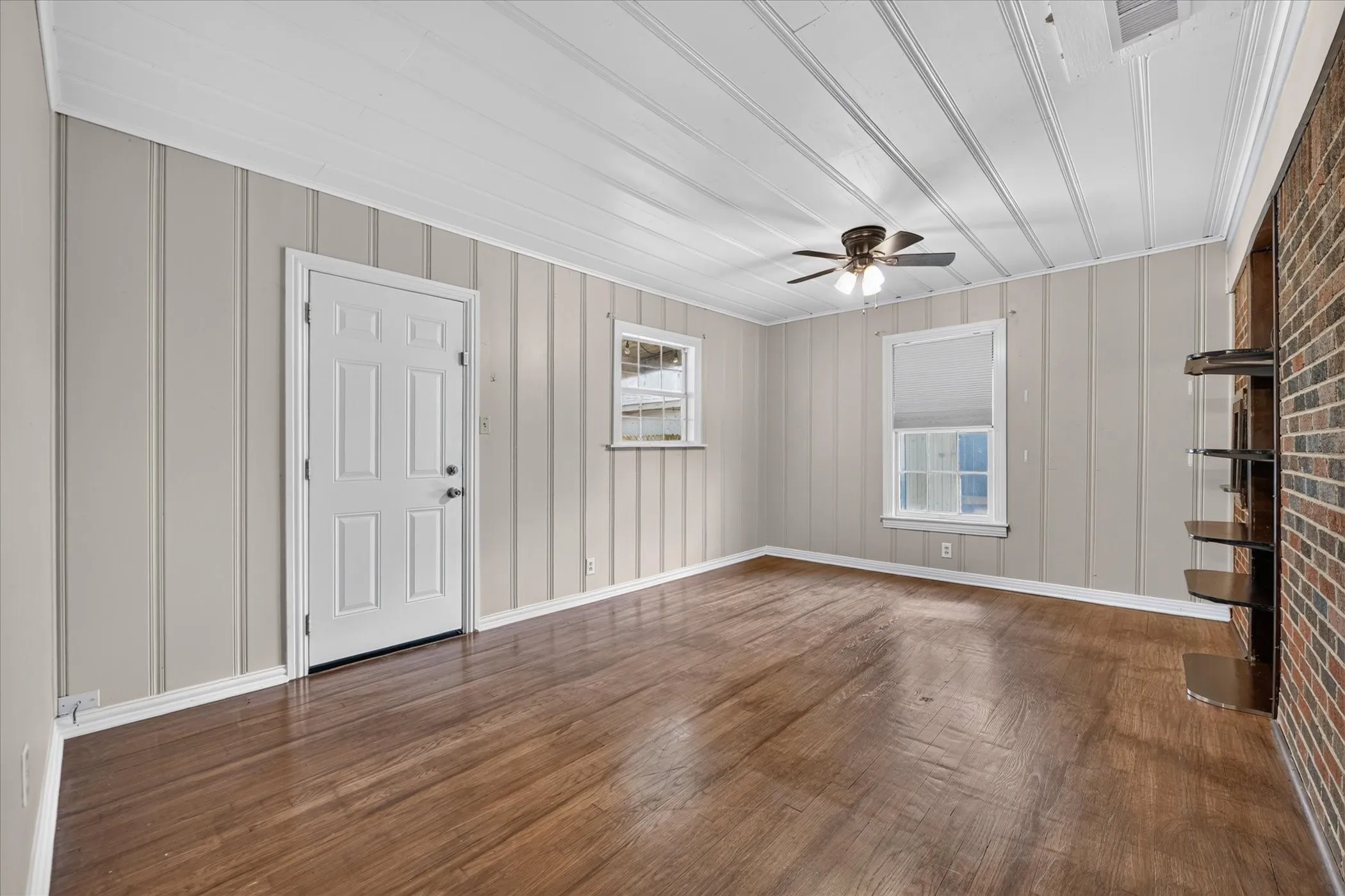 Wood-finish flooring, vertical paneling, brick accent wall, ceiling fan, and built-in shelving