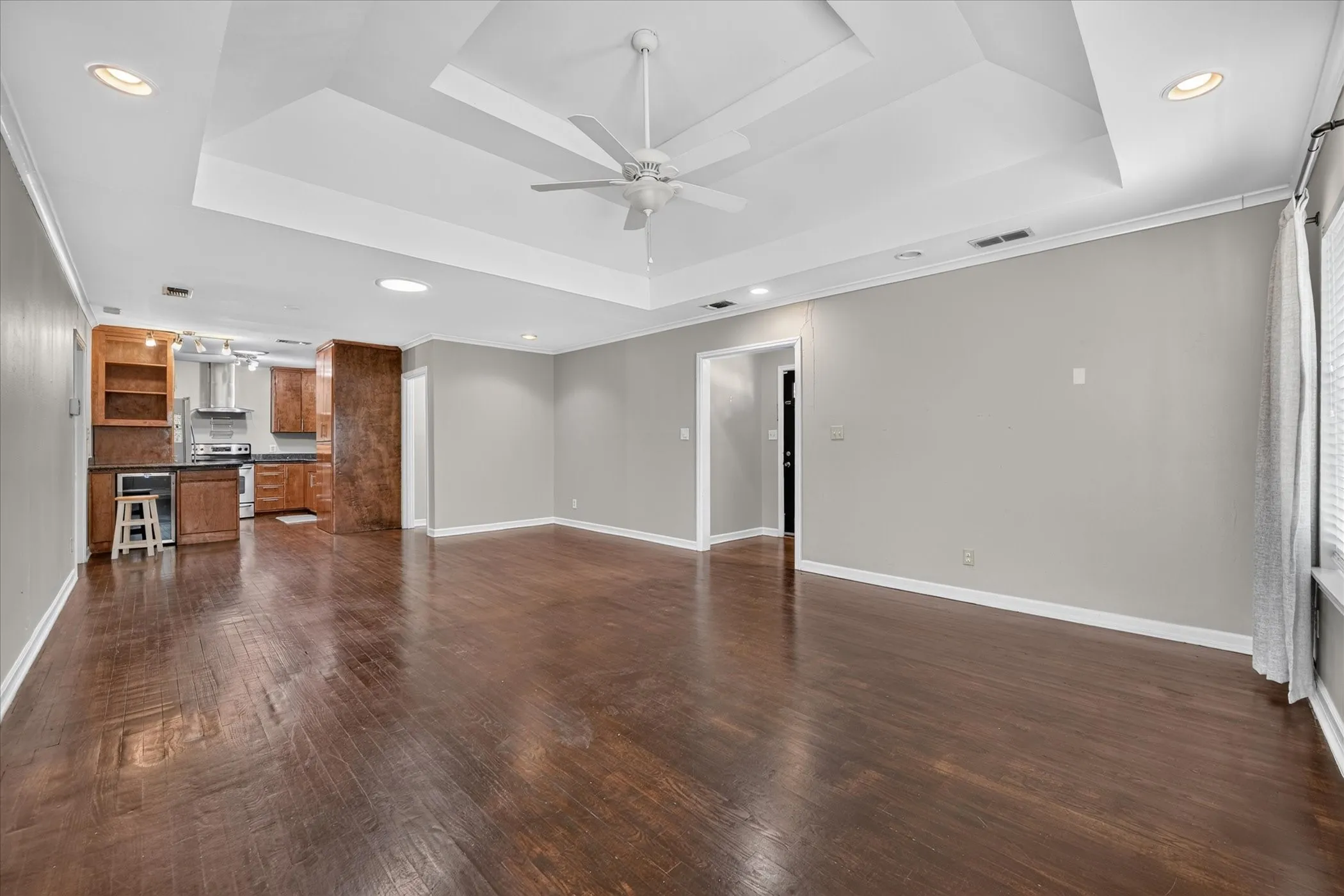 Spacious room featuring wood-finish flooring, a tray ceiling with a ceiling fan, recessed lighting, and a partial kitchen with wood cabinetry and stainless steel appliances