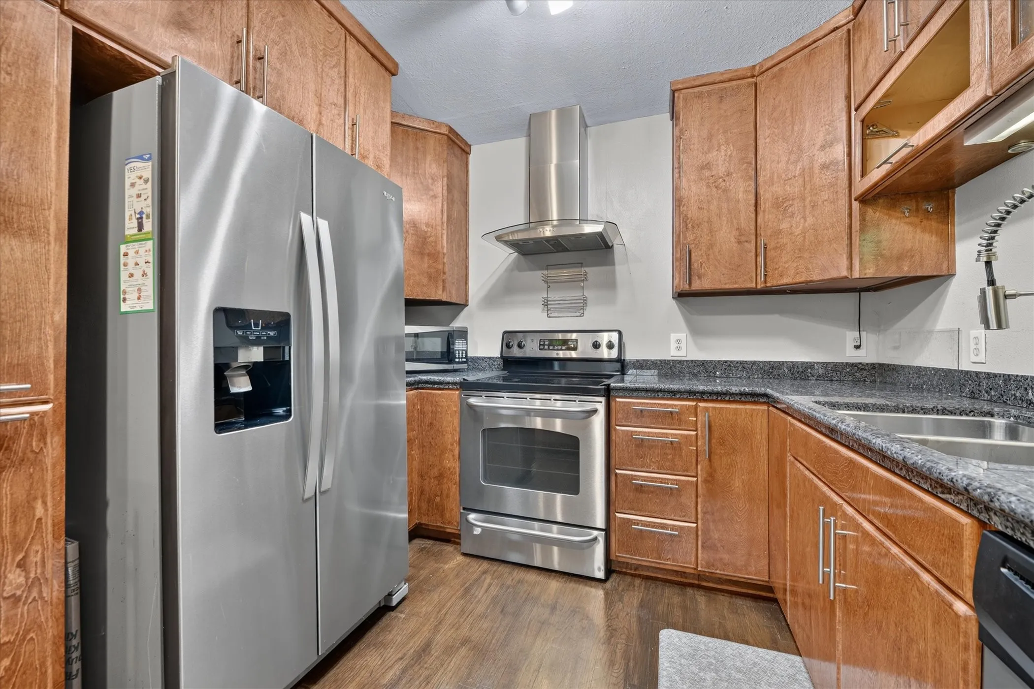 Full kitchen featuring stainless steel appliances, wood-finish cabinetry, dark countertops, and a modern range hood