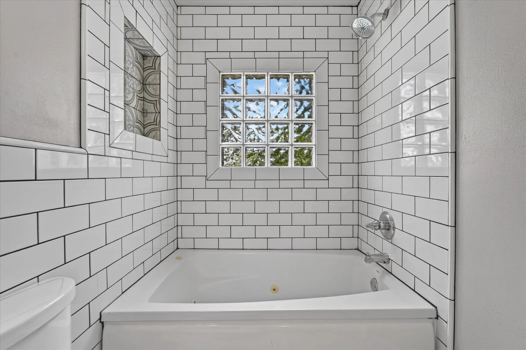 White subway tile surround, built-in tub with chrome fixtures, glass block window, and decorative inset shelf