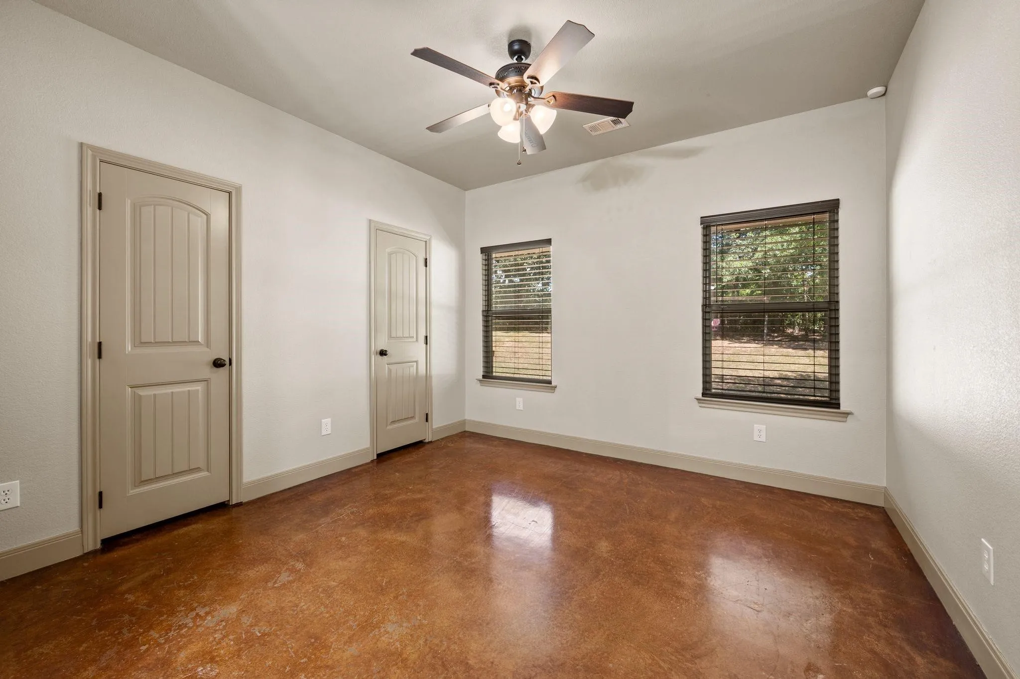 Stained concrete flooring, two windows with blinds, two paneled doors, and a ceiling fan with integrated lighting