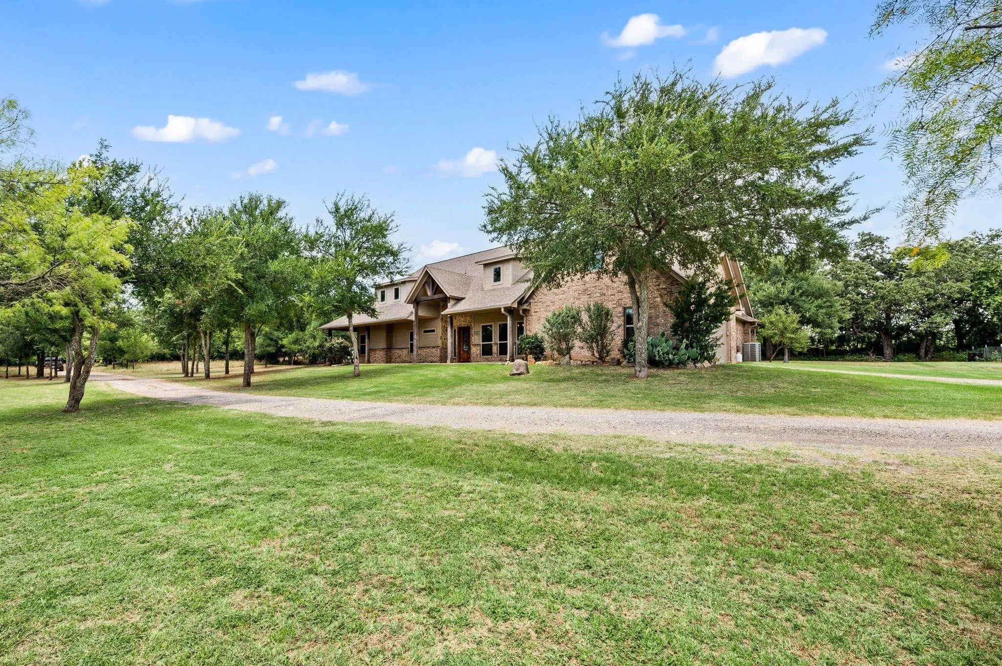 Brick exterior home with multiple gables and dormer windows, featuring a gravel driveway, mature trees, and a grassy expanse
