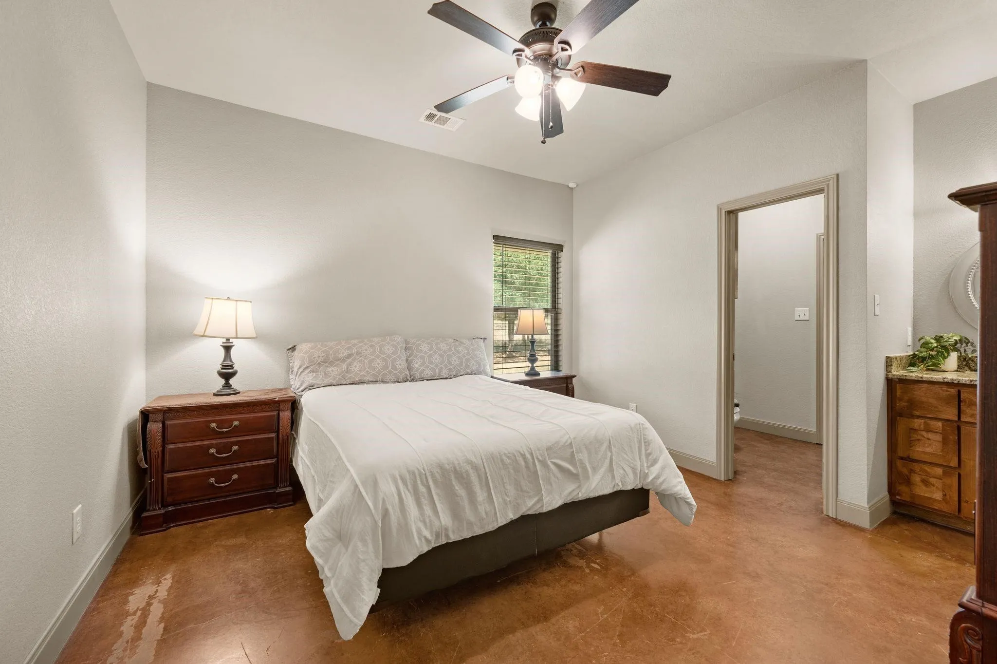Smooth concrete flooring, ceiling fan with integrated lighting, window with horizontal blinds, built-in vanity with stone countertop, and wood-finish cabinetry