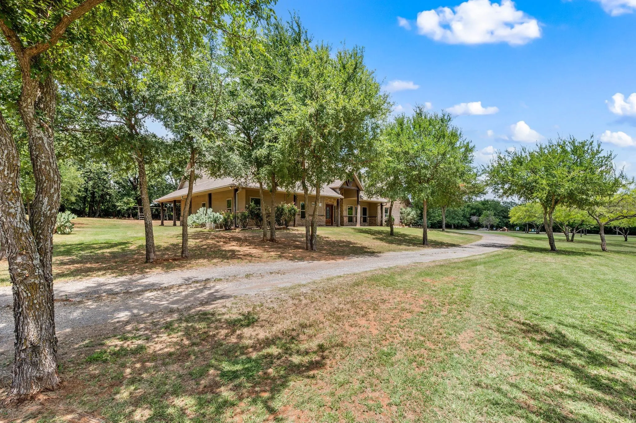 Expansive property featuring a gravel driveway, mature trees, a covered porch, and a pitched roofline