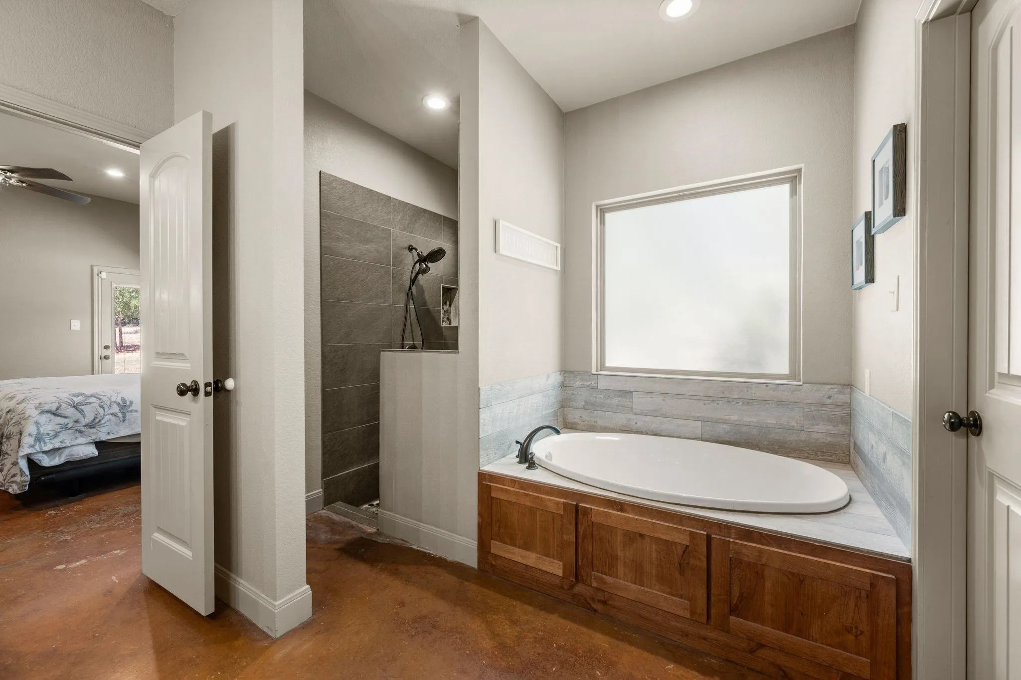 Soaking tub with a wood-finish surround, large frosted window, and an adjacent walk-in shower featuring dark tile