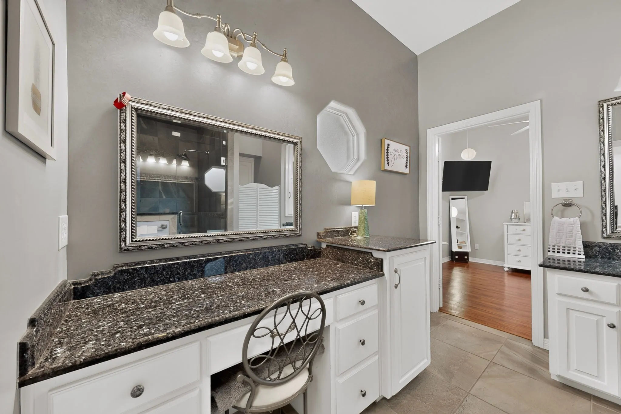 Vaulted ceiling, white cabinetry with dark hardware, stone countertops, vanity mirror with ornamental frame, and tile flooring