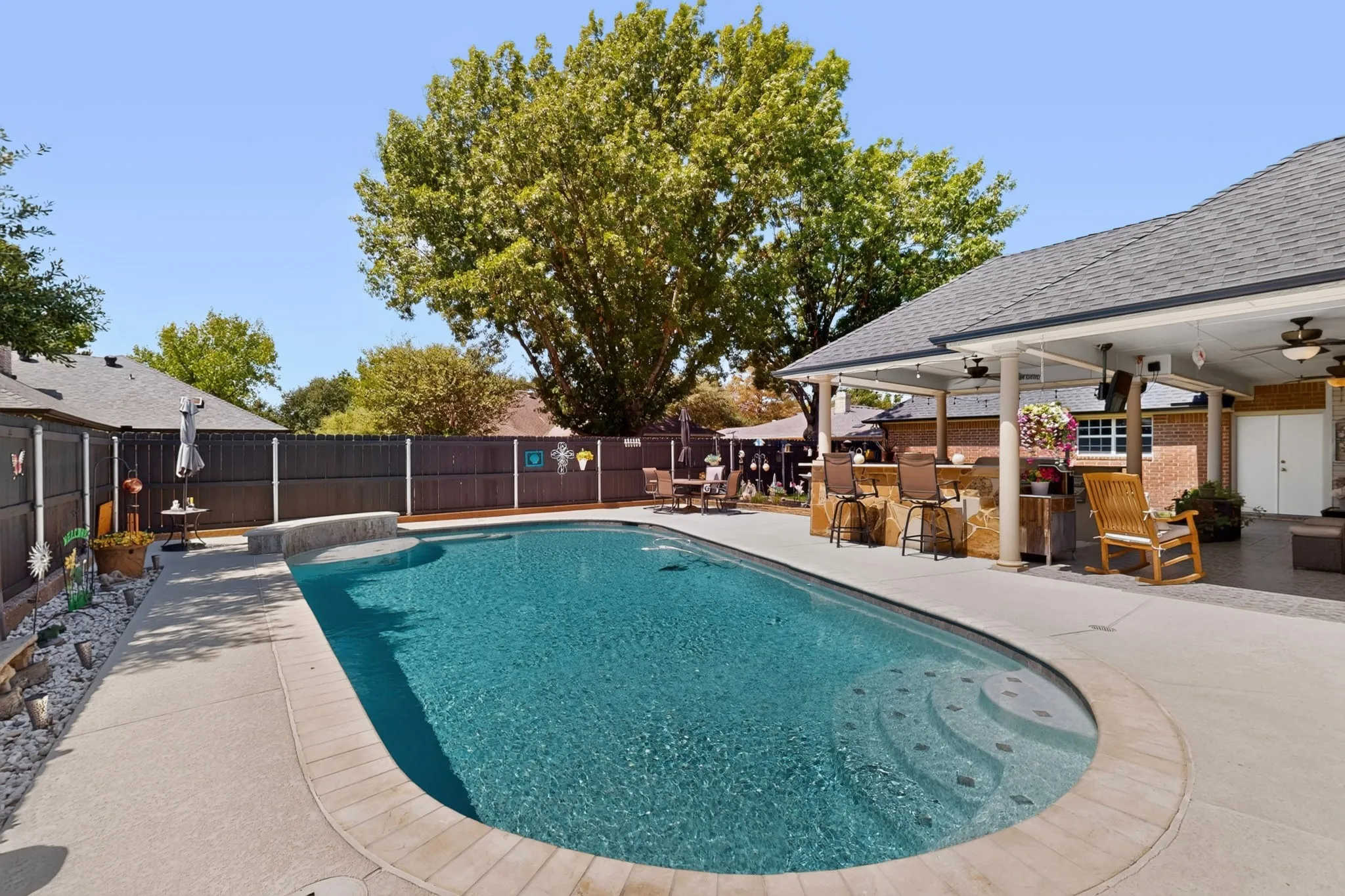 Inground swimming pool with integrated steps, expansive concrete decking, and an outdoor covered patio featuring a built-in bar