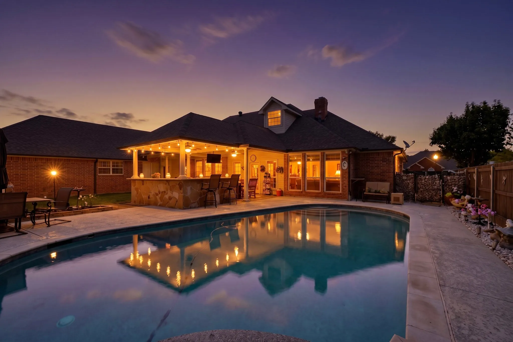 Curved swimming pool, stone-clad outdoor bar, covered patio, multiple large windows, and brick exterior