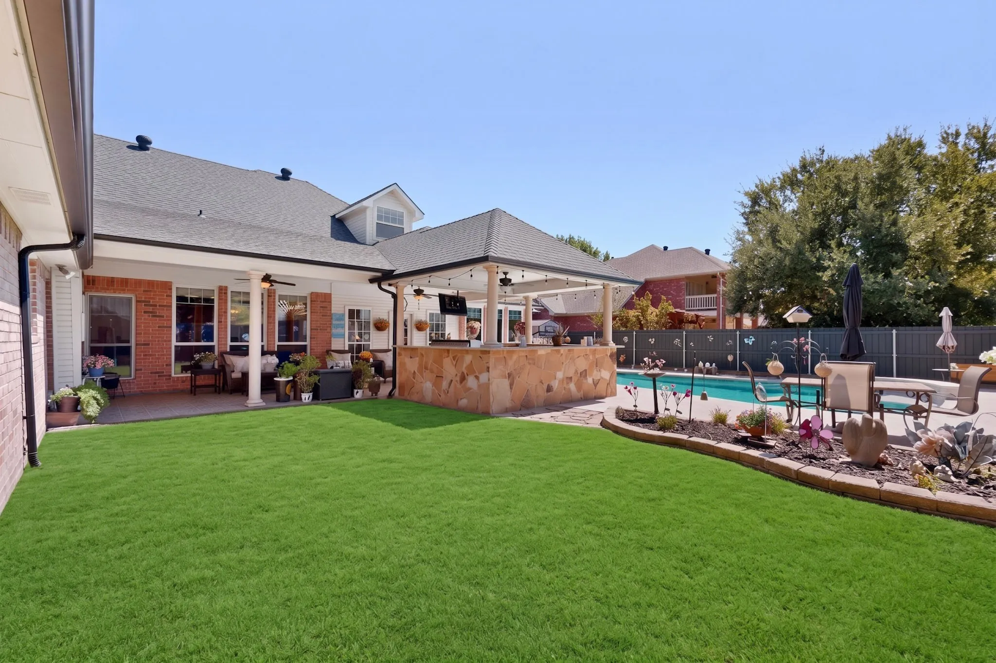 Expansive backyard featuring a manicured lawn, a covered patio, an outdoor kitchen with a stone-clad bar, and a swimming pool with a surrounding paved deck