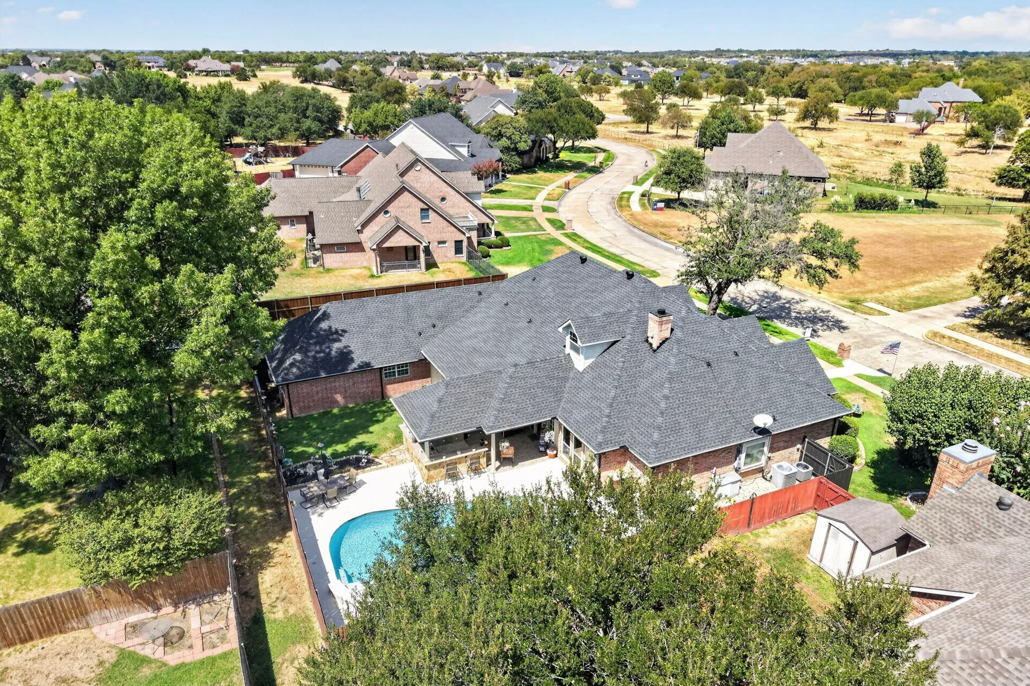 Expansive backyard featuring a curved swimming pool, a wood fence, and a brick exterior home with a dark shingle roof