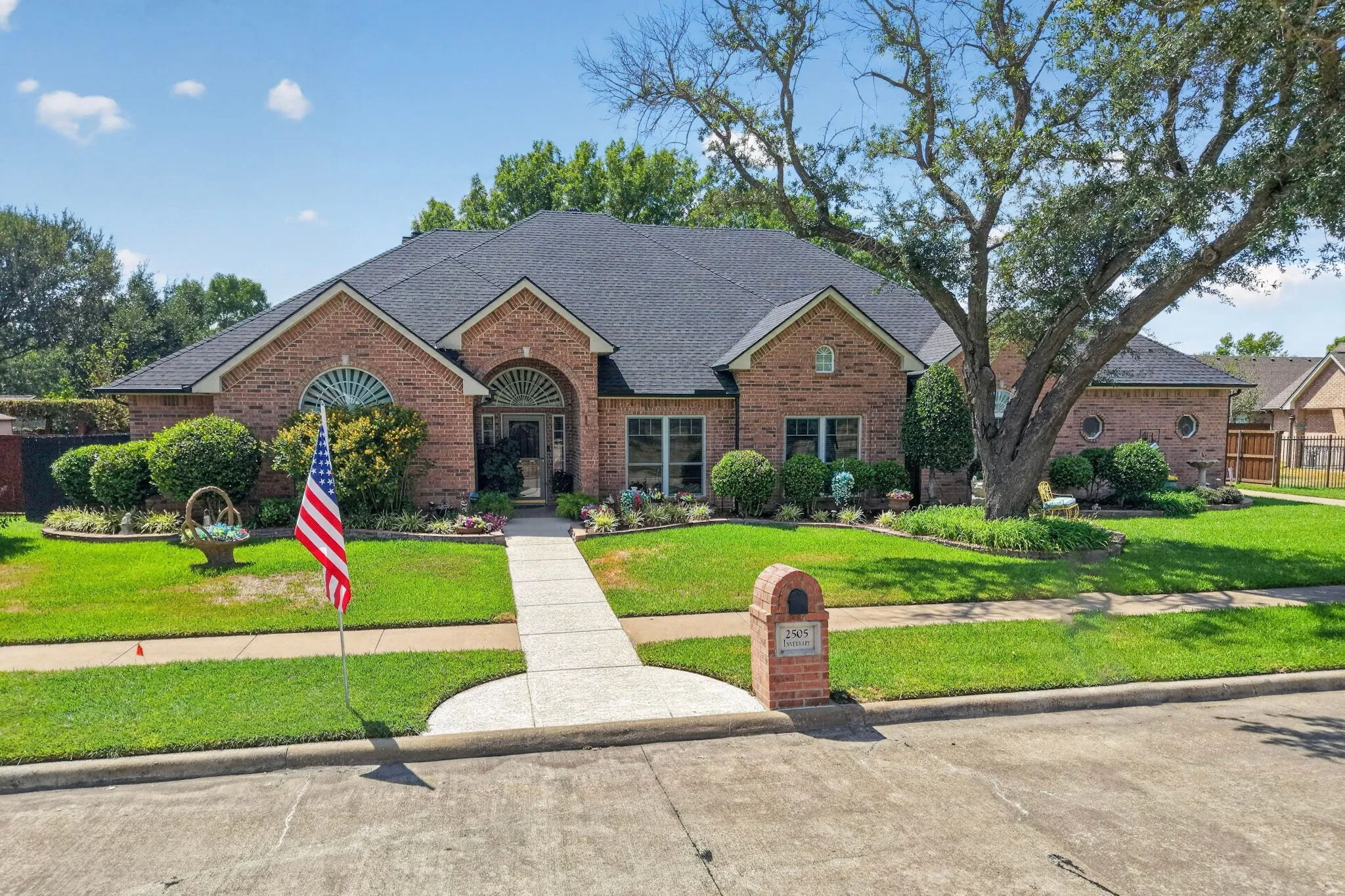 Brick exterior with arched windows, dark shingle roof, established landscaping, and a concrete walkway