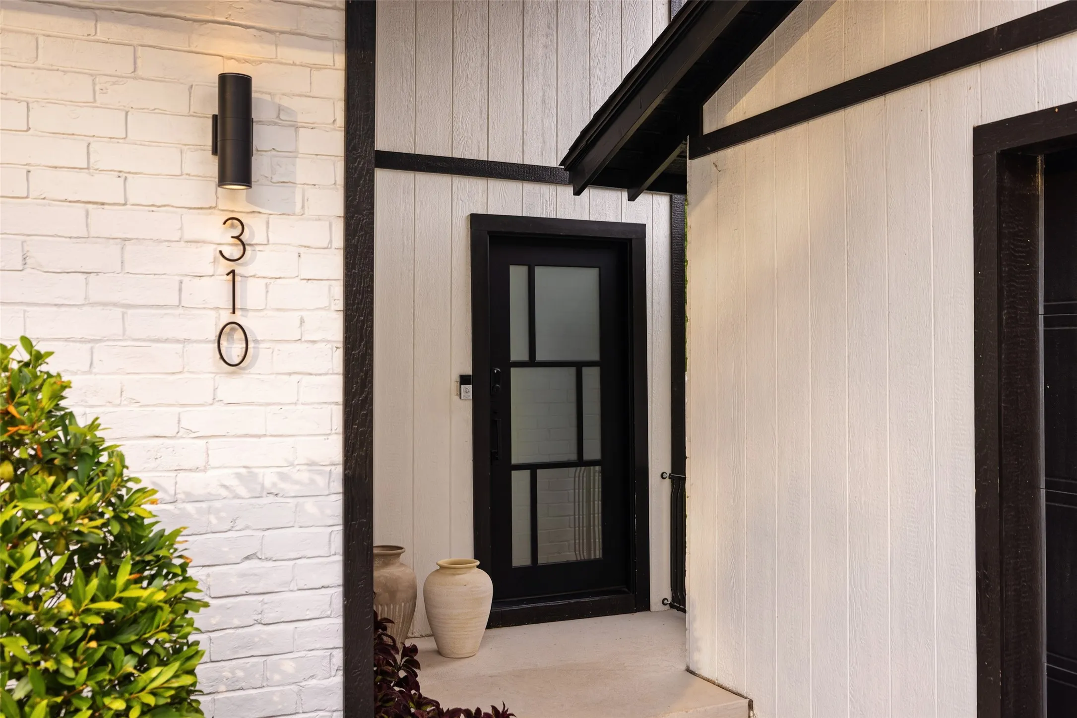 White painted brick facade with a black cylindrical outdoor light fixture, contrasting black window trim, and a black paneled door with frosted glass inserts