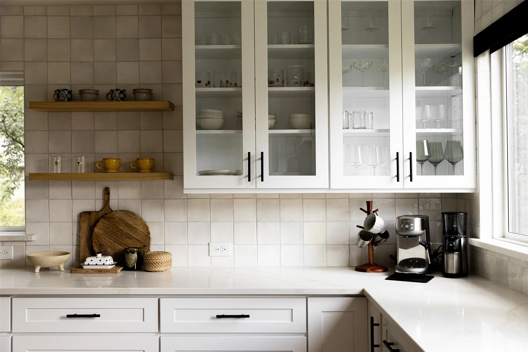 White shaker cabinetry, glass-front upper cabinets, light-toned square tile backsplash, floating wood shelves, and light-toned countertops