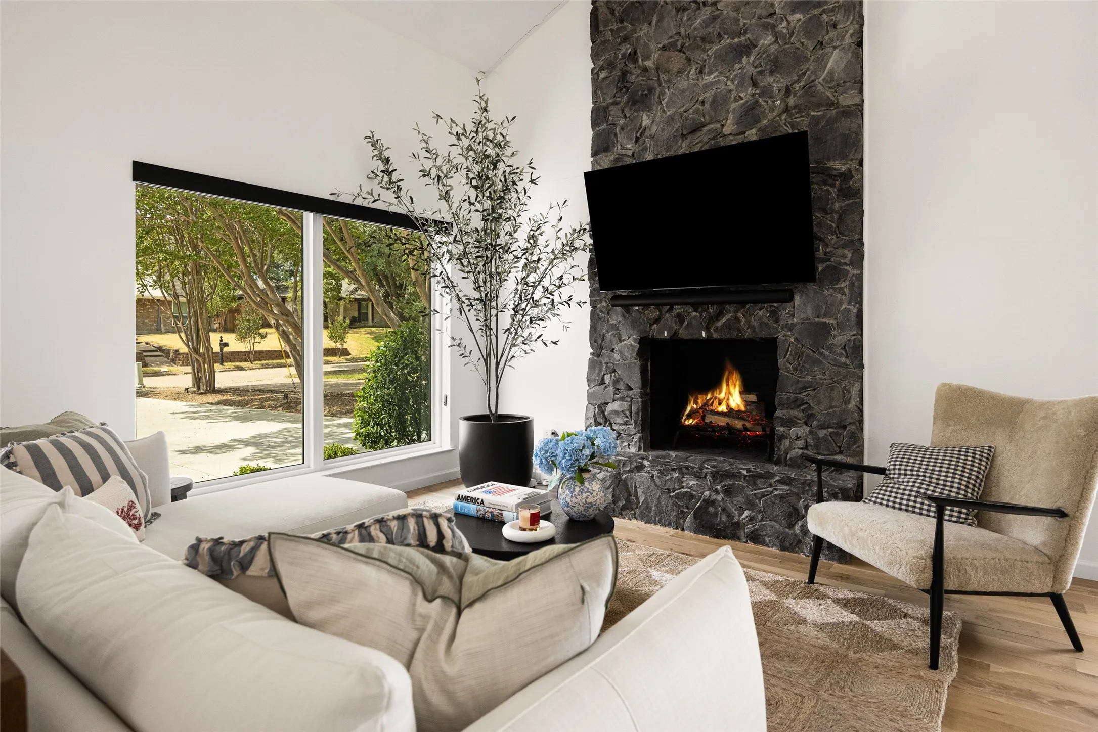 Expansive floor-to-ceiling windows, stone-clad fireplace with hearth, and light wood-finish flooring