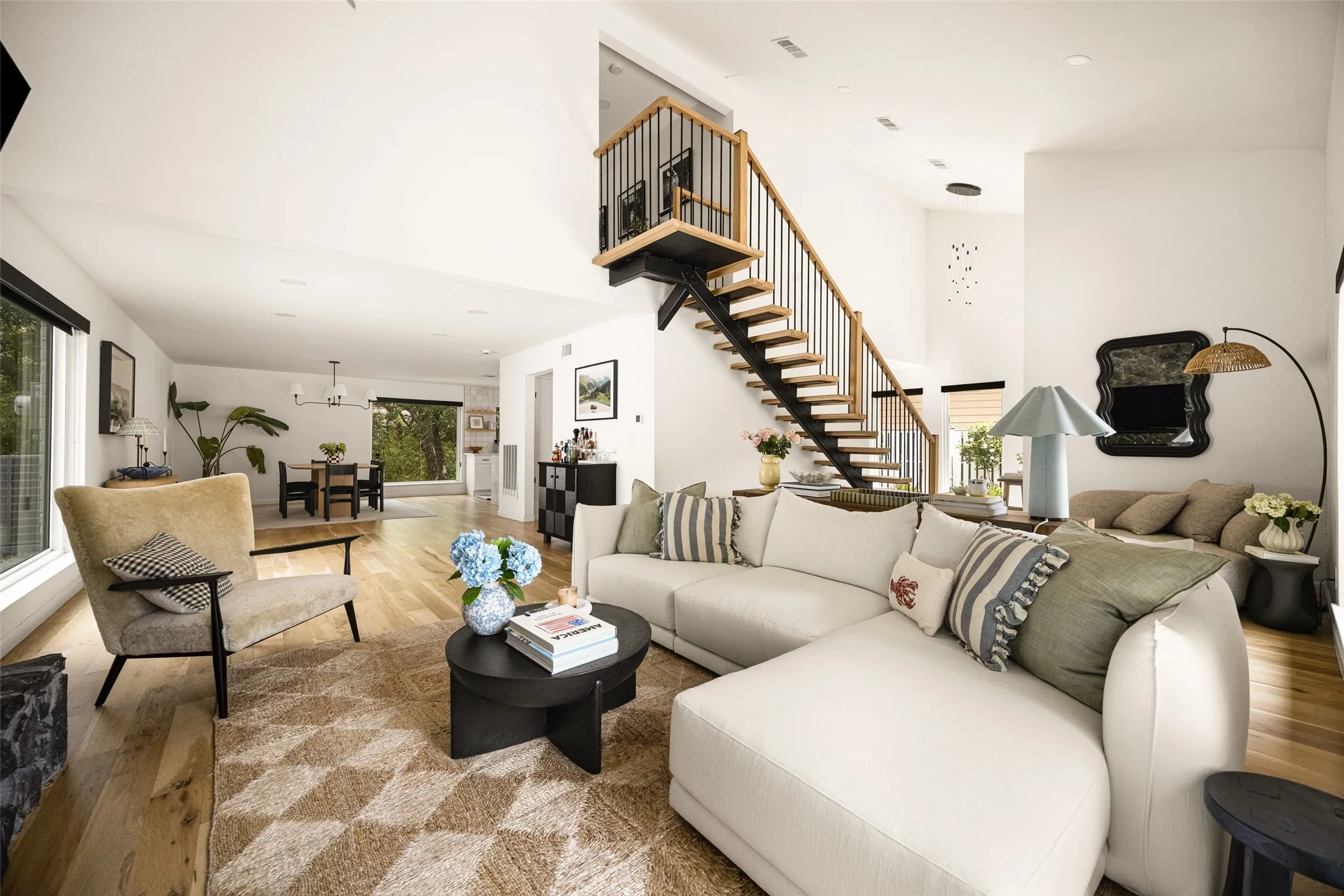 Open-concept living area with wood-finish flooring, a contemporary staircase featuring black spindles and wood handrails, and large windows providing abundant light