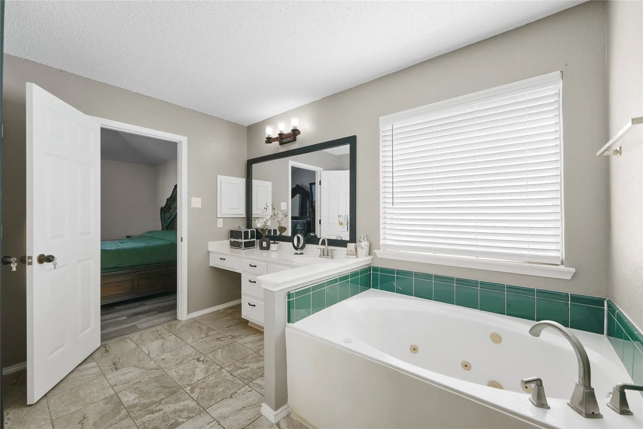 Jetted soaking tub with green tile surround, dual vanity with built-in storage, and tile flooring