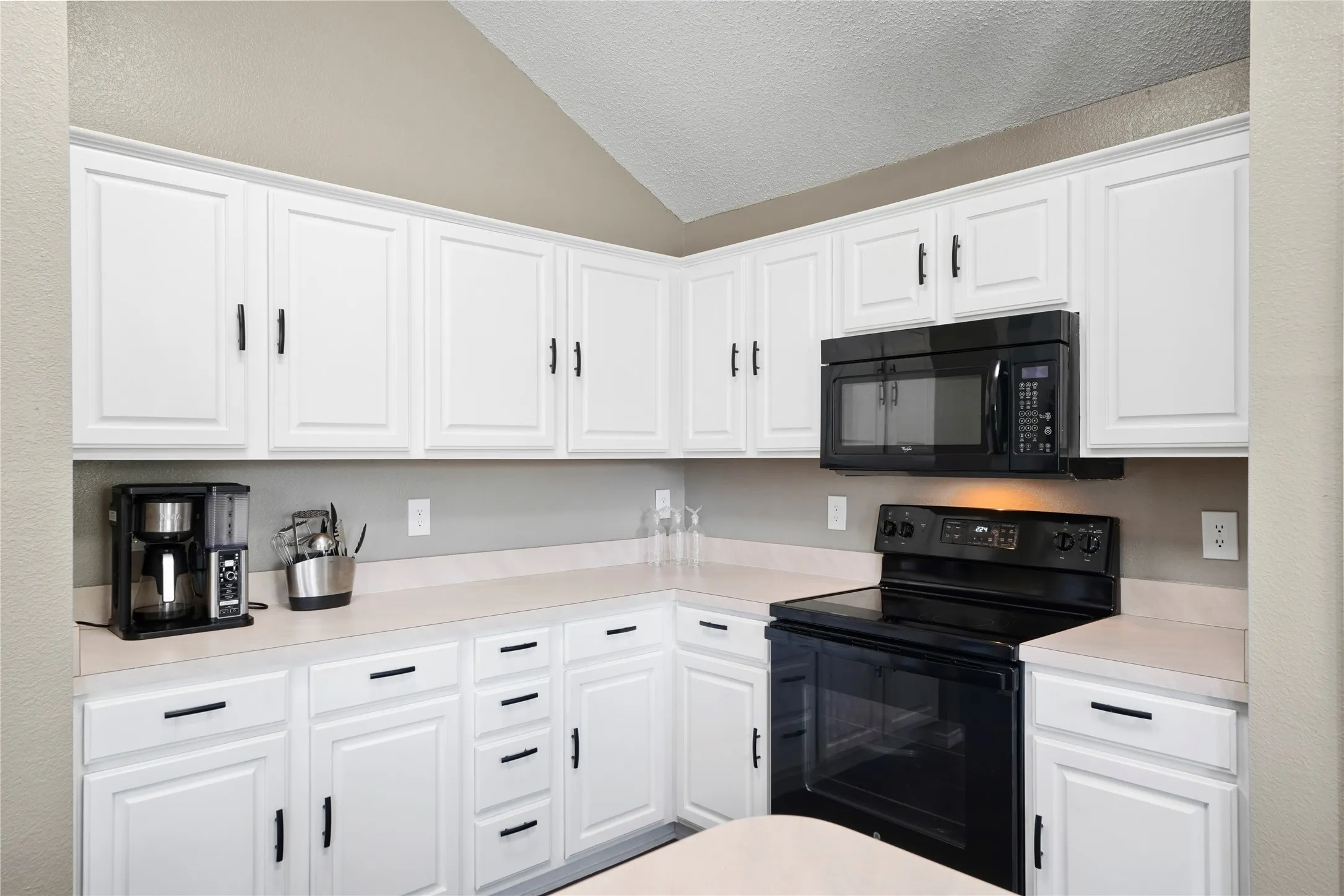 Vaulted ceiling, white shaker-style cabinetry, black hardware, black microwave, and black electric range