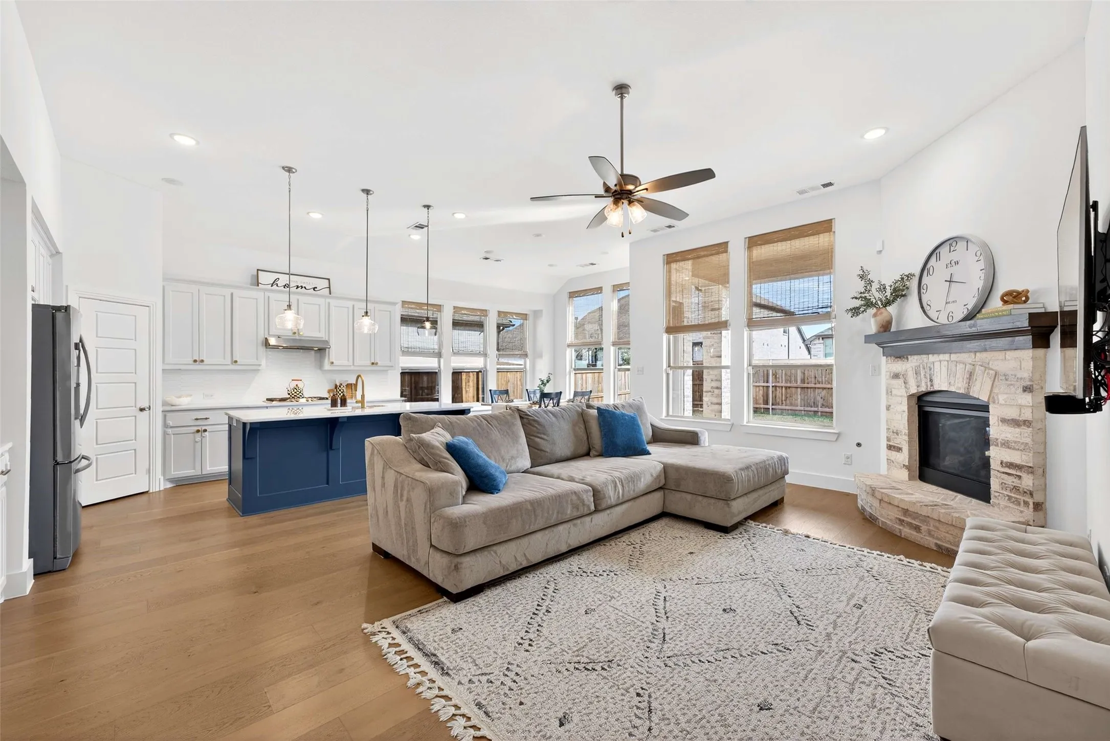 Open-concept living space featuring wood-finish flooring, a kitchen island with pendant lighting, a ceiling fan, and a fireplace with a stone surround