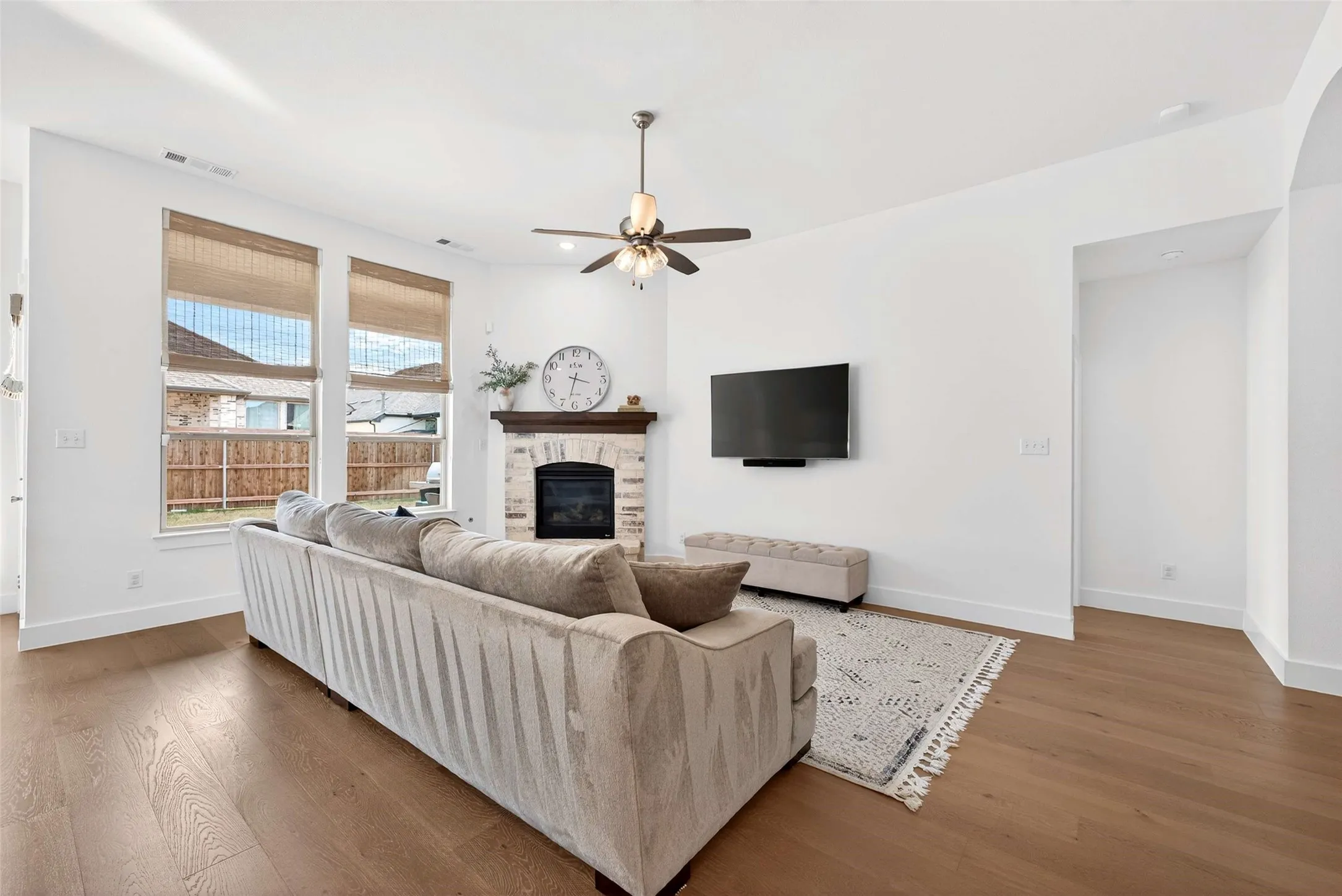 Hardwood-style flooring, white walls, stone-clad fireplace with wood mantel, ceiling fan, and arched entryway