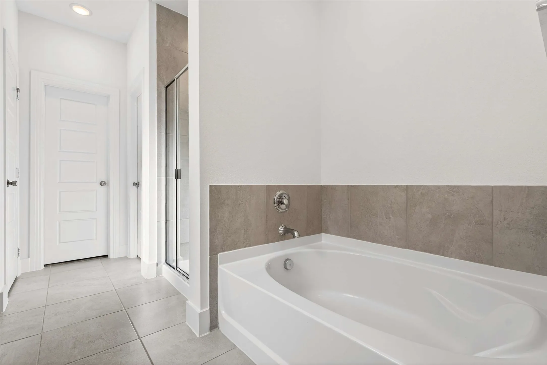 White soaking tub with chrome fixtures, floor-to-ceiling tiled shower enclosure, and light gray floor tiling