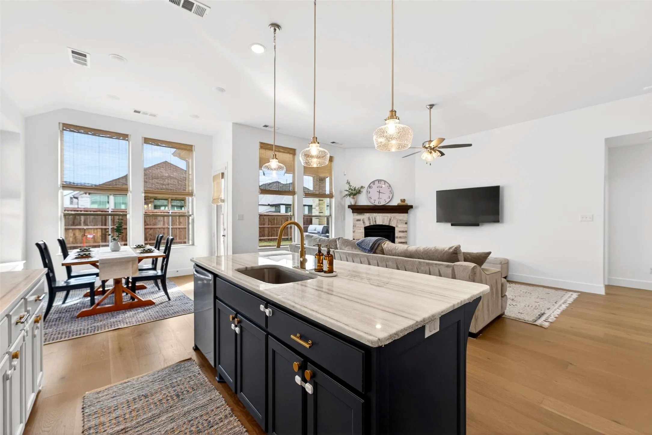 Open-concept living area with wood-finish flooring, a kitchen island with a built-in sink, pendant lighting, and a stone-clad fireplace