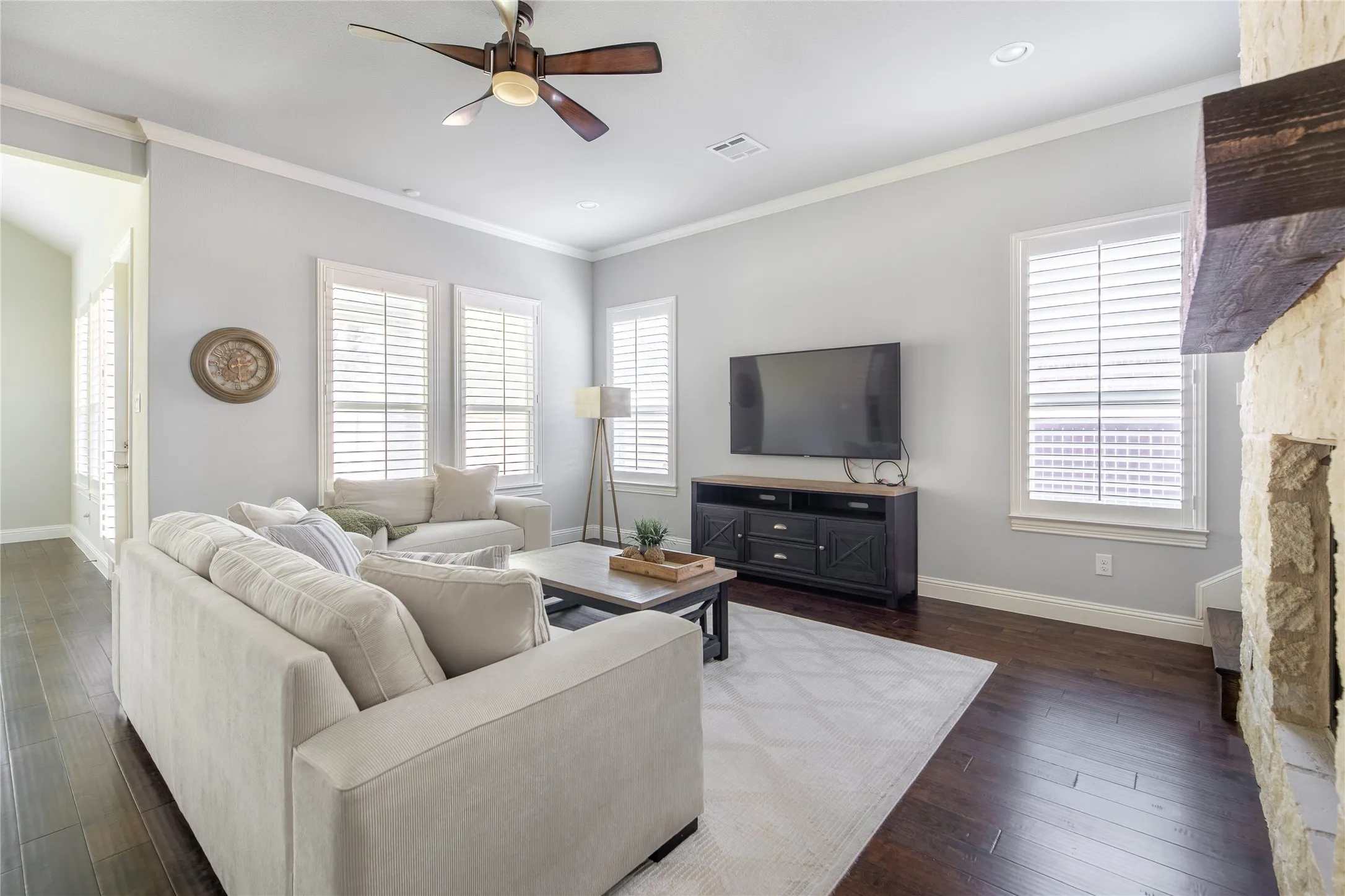 Wood-finish flooring, white plantation shutters, stone fireplace with a wood mantel, and a ceiling fan with integrated lighting
