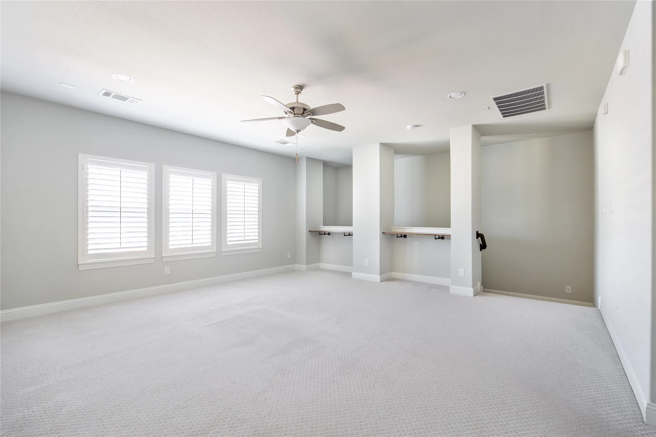 Spacious room featuring light-colored carpeting, three windows with white plantation shutters, a ceiling fan, and built-in desk nooks