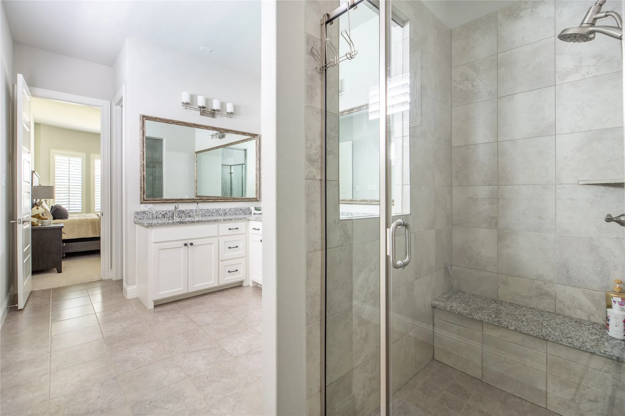 Enclosed shower with tiled walls and built-in bench, double vanity with granite countertop, large framed mirror, and tiled flooring