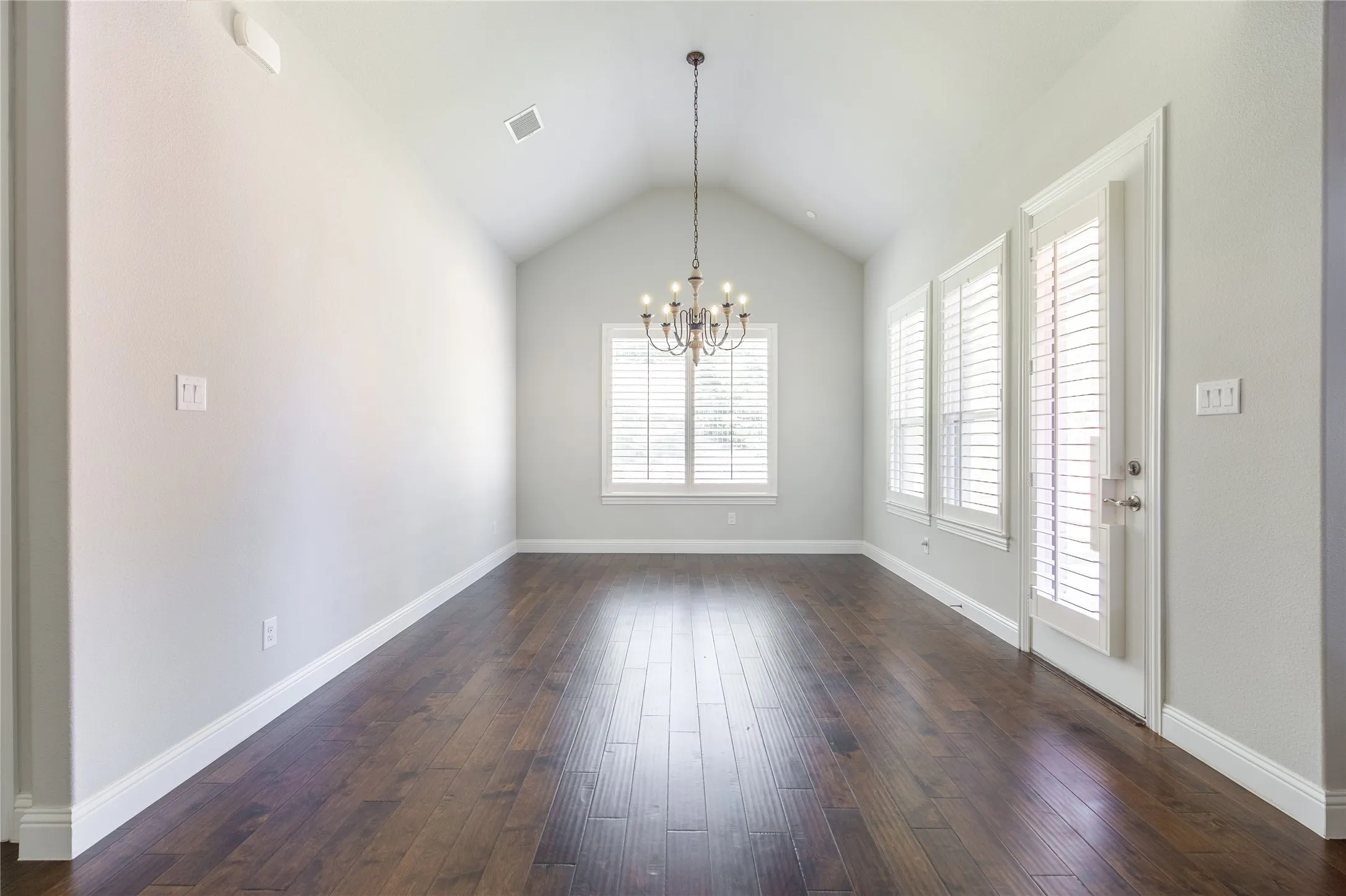 Vaulted ceiling with an elegant chandelier, wood-finish flooring, multiple windows with plantation shutters, and a white paneled door
