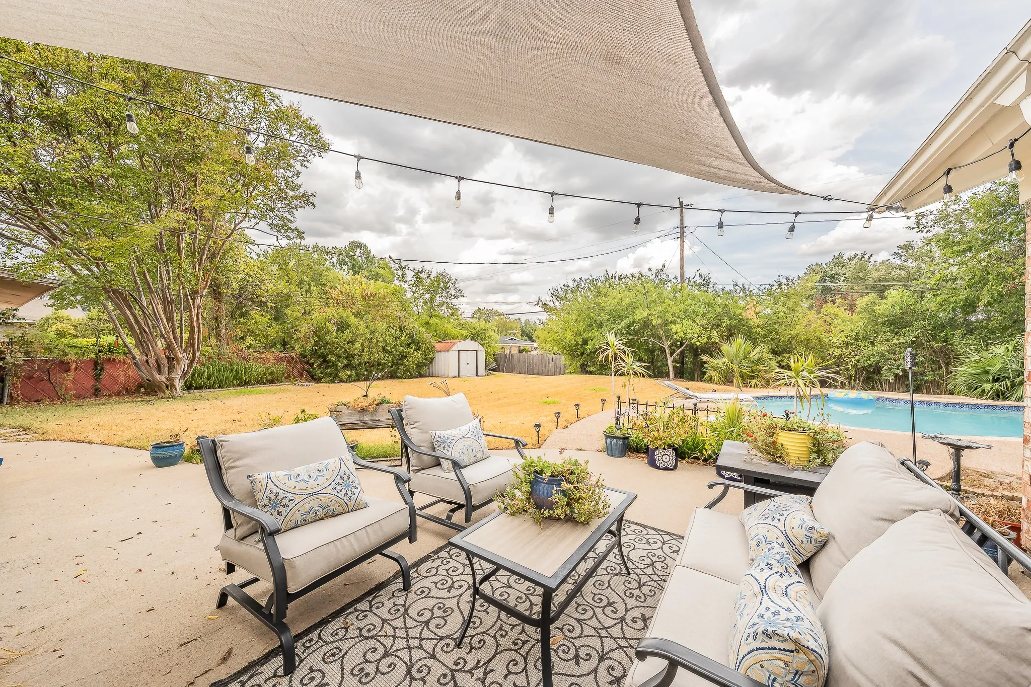 Concrete patio with a shade sail, string lighting, inground pool, storage shed, and mature trees