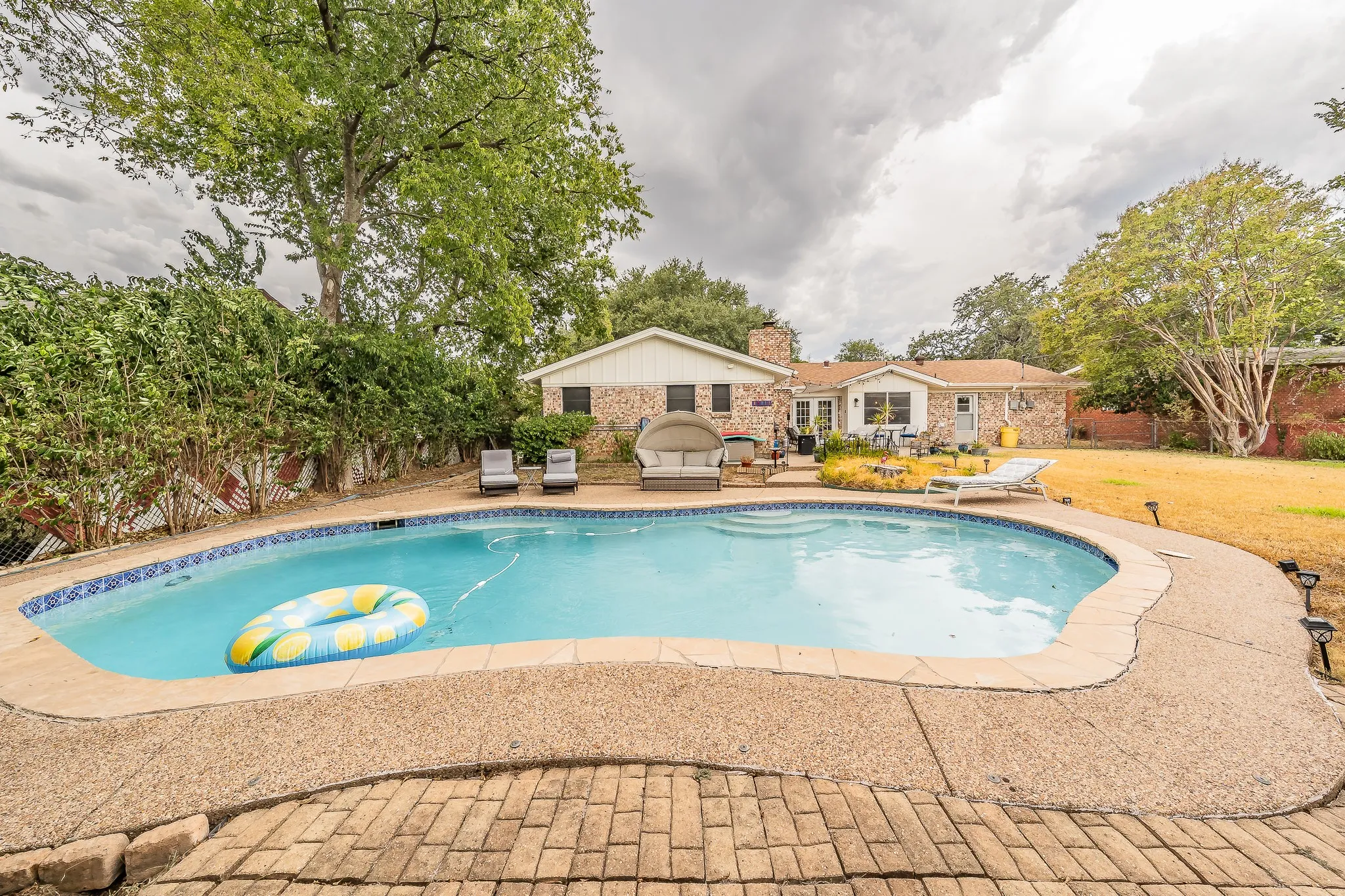 Outdoor swimming pool with a stone coping and brick paver patio, surrounded by mature landscaping and a partial brick exterior residence
