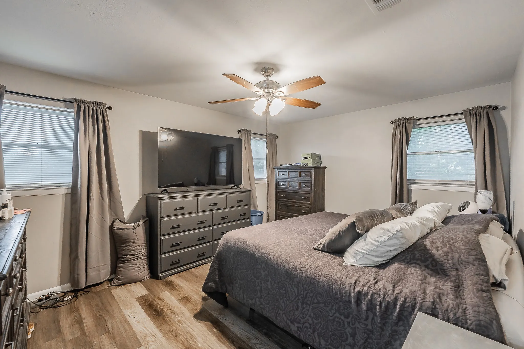 Spacious primary bedroom featuring wood-finish flooring, dual windows with thermal-lined drapery, ceiling fan with integrated lighting, and coordinating wood furniture pieces including dresser and media console