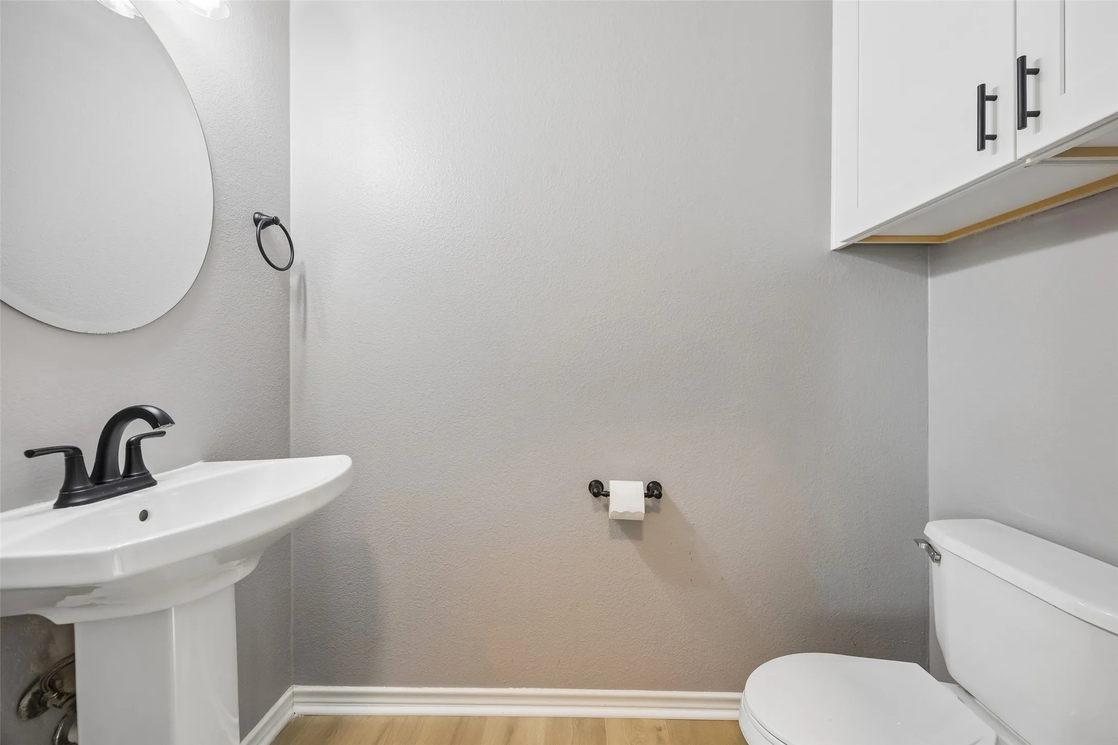 Pedestal sink with a matte black faucet, wall-mounted circular mirror, white cabinetry with black hardware, and wood-finish flooring