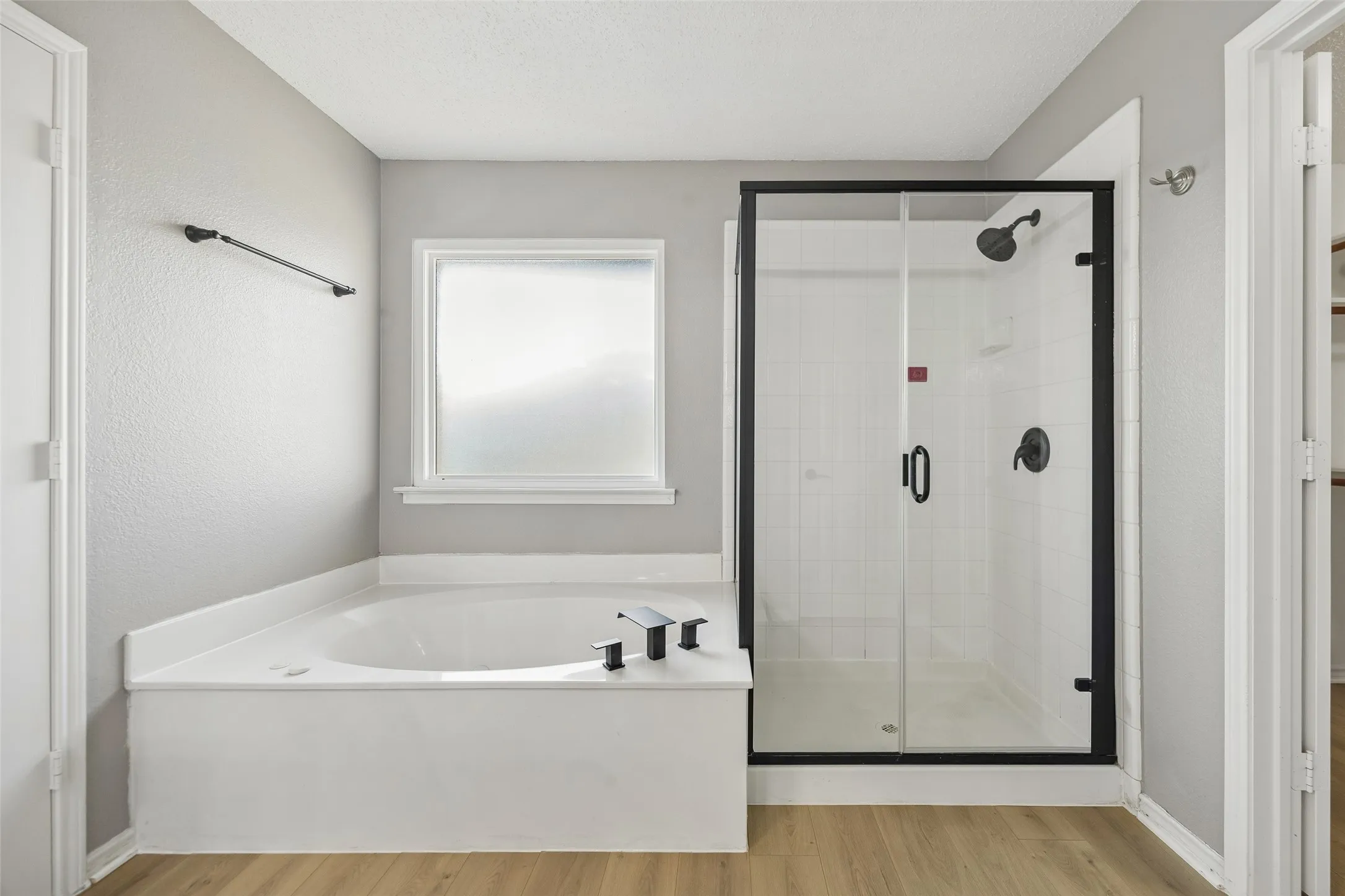 Soaking tub with black fixtures, enclosed shower with black frame, white subway tile shower surround, and wood-finish flooring