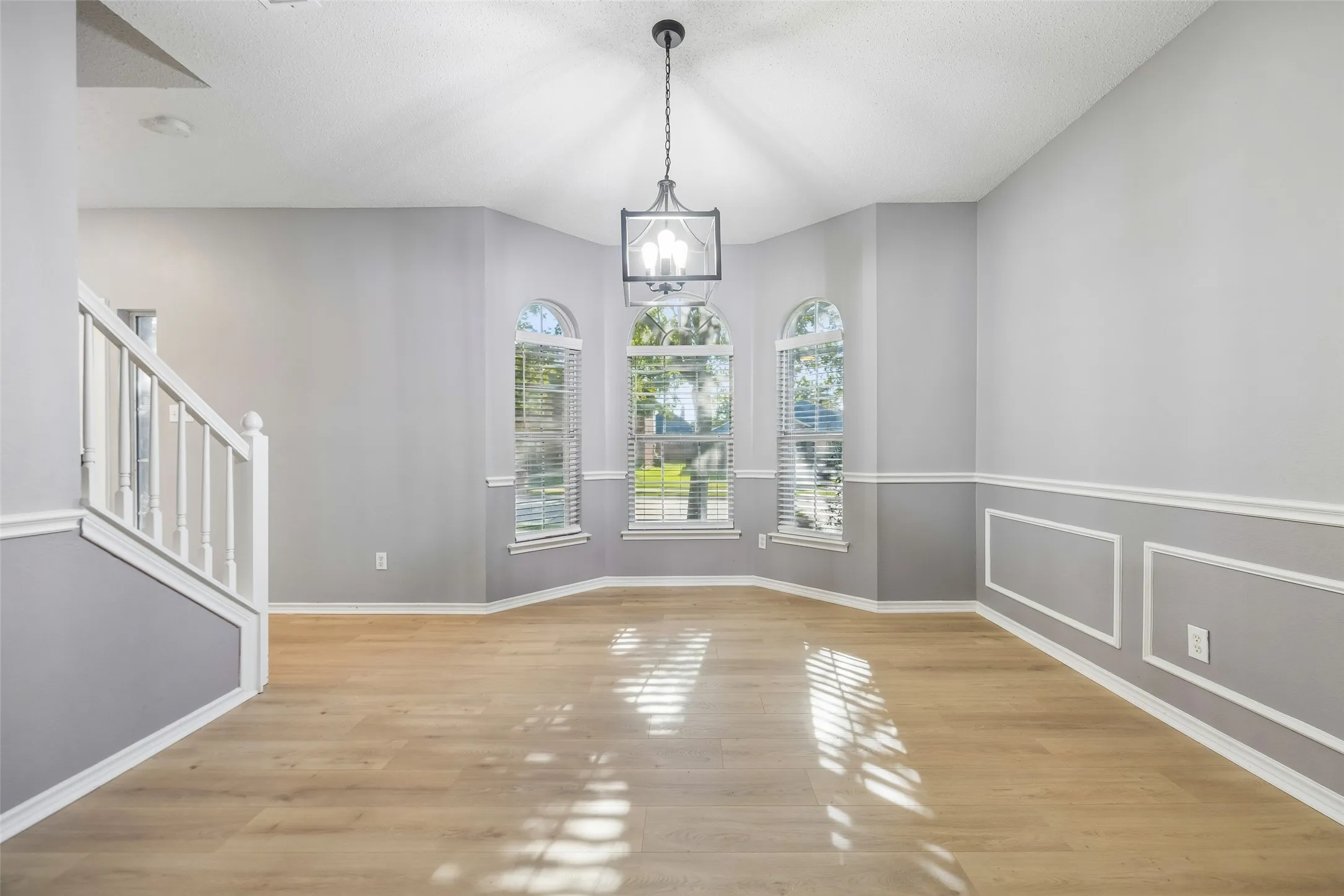 Vaulted ceiling featuring a decorative chandelier, multiple arched windows with blinds, wainscoting details, wood-finish flooring, and staircase with white banister