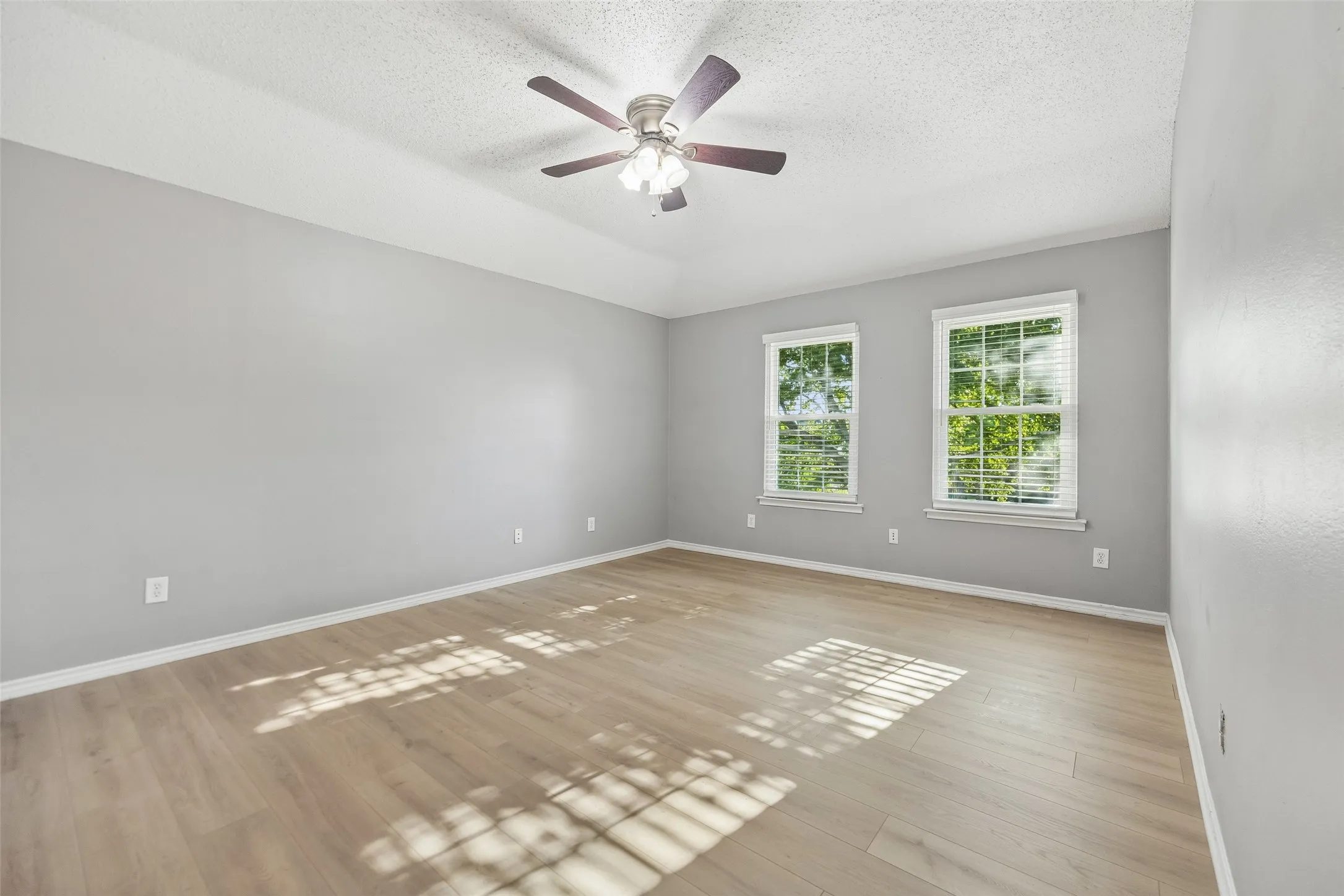 Spacious room featuring wood-finish flooring, light gray walls, two windows with blinds, and a ceiling fan