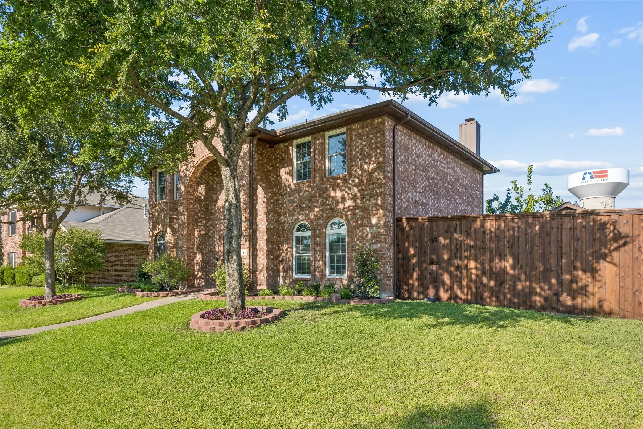 Brick exterior with arched entryway, multiple windows, established trees, and a privacy fence