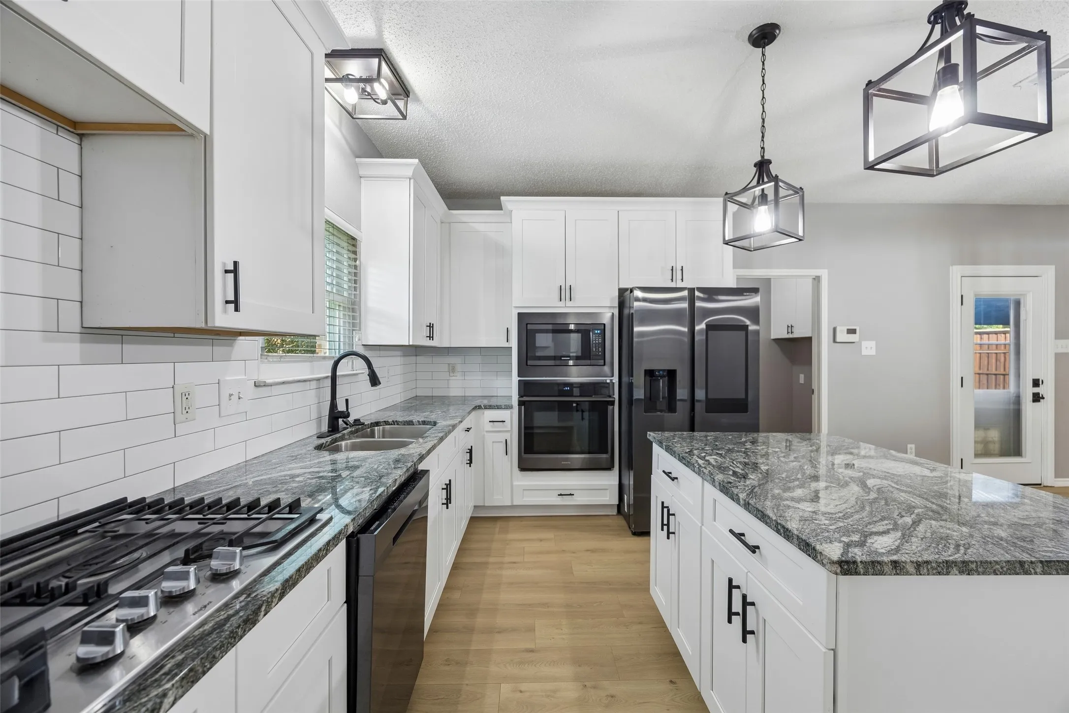 White cabinetry with matte black hardware, integrated stainless steel appliances, wood-finish flooring, and a large central island with a patterned countertop