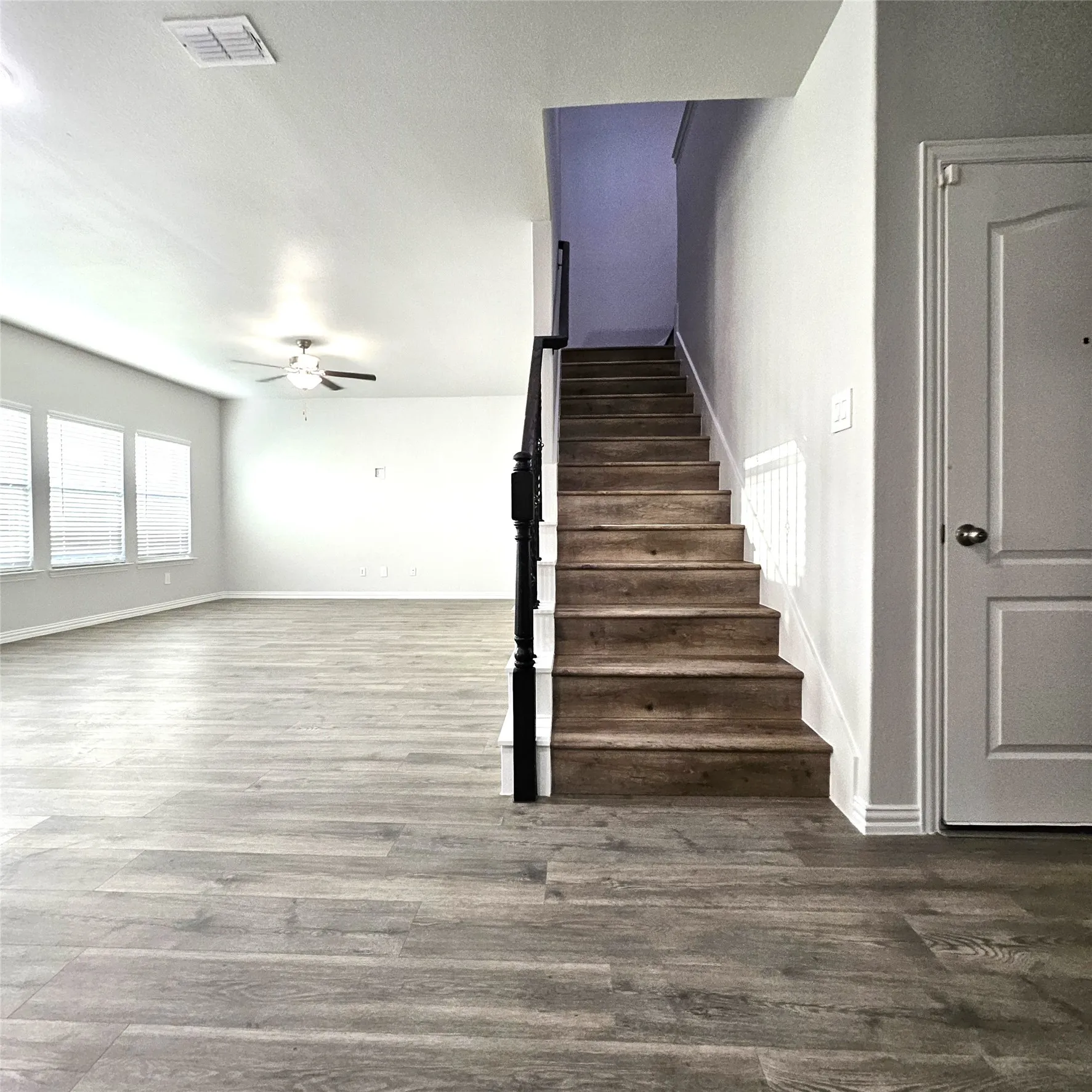 Spacious living area featuring wood-finish flooring, multiple windows with blinds, a ceiling fan, and a staircase with a dark wood-finish handrail