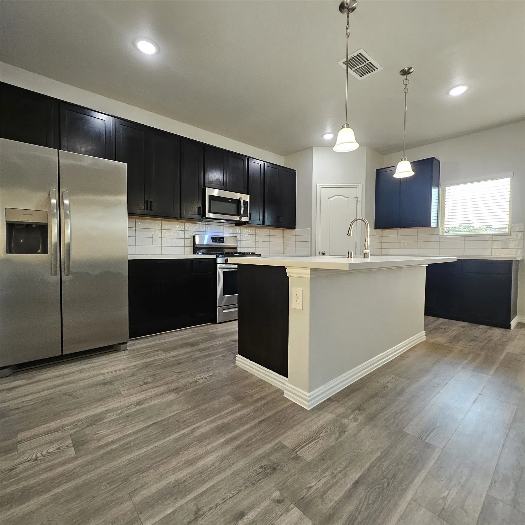 Kitchen with wood-finish flooring, dark cabinetry, stainless steel appliances, a white subway tile backsplash, and an island with a built-in sink