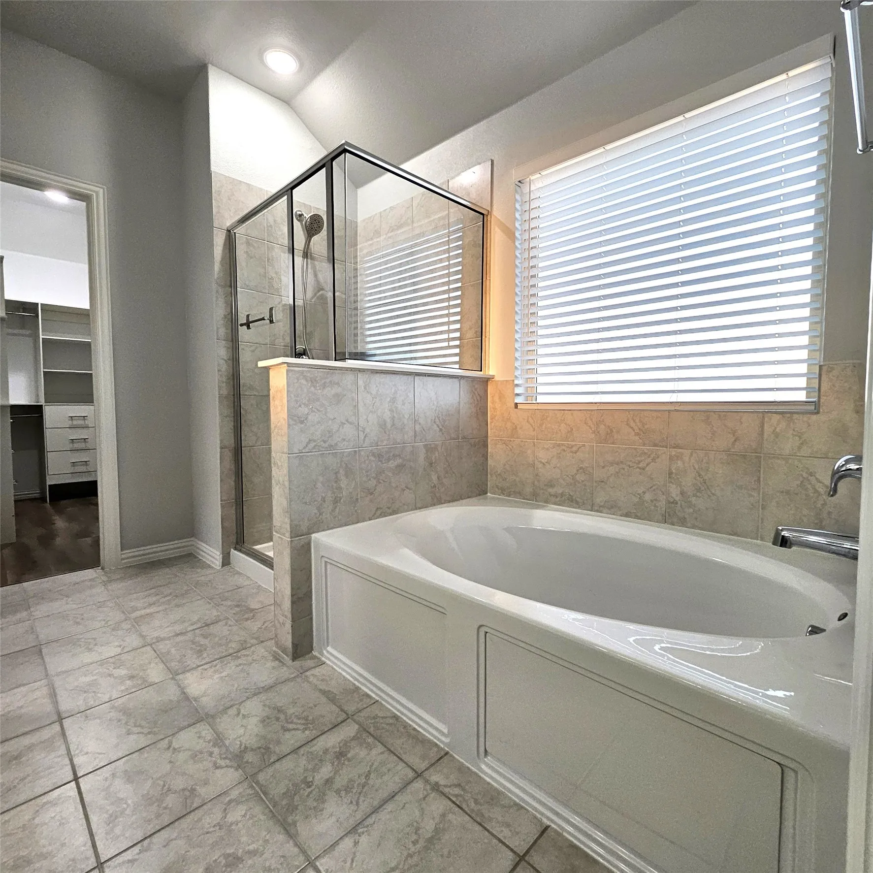 Tile flooring, built-in soaking tub, glass-enclosed shower, tiled half-wall, and a window with blinds