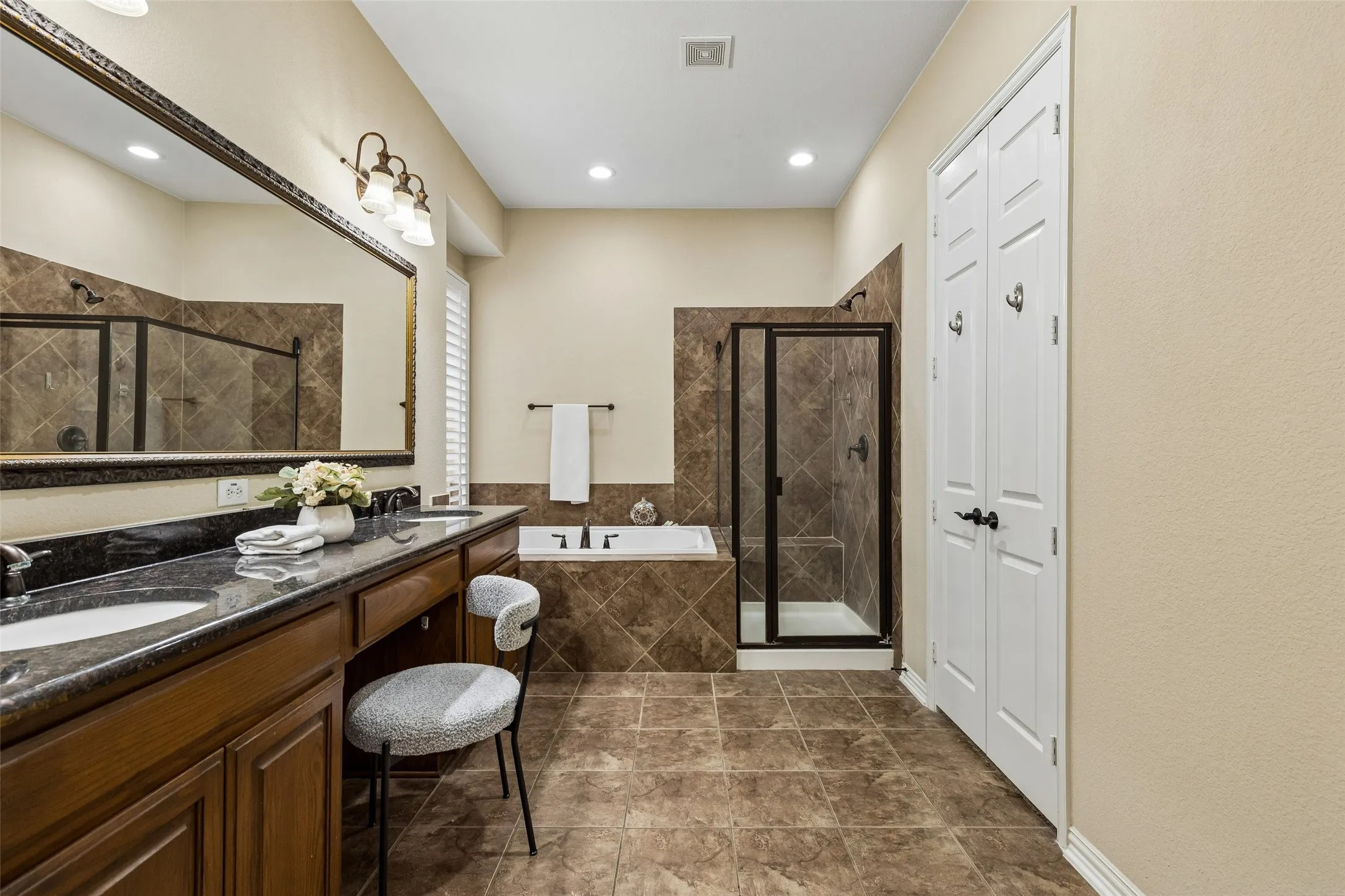 Dual vanity with integrated sinks, built-in soaking tub, separate glass-enclosed shower, wood-finish cabinetry, and tiled flooring