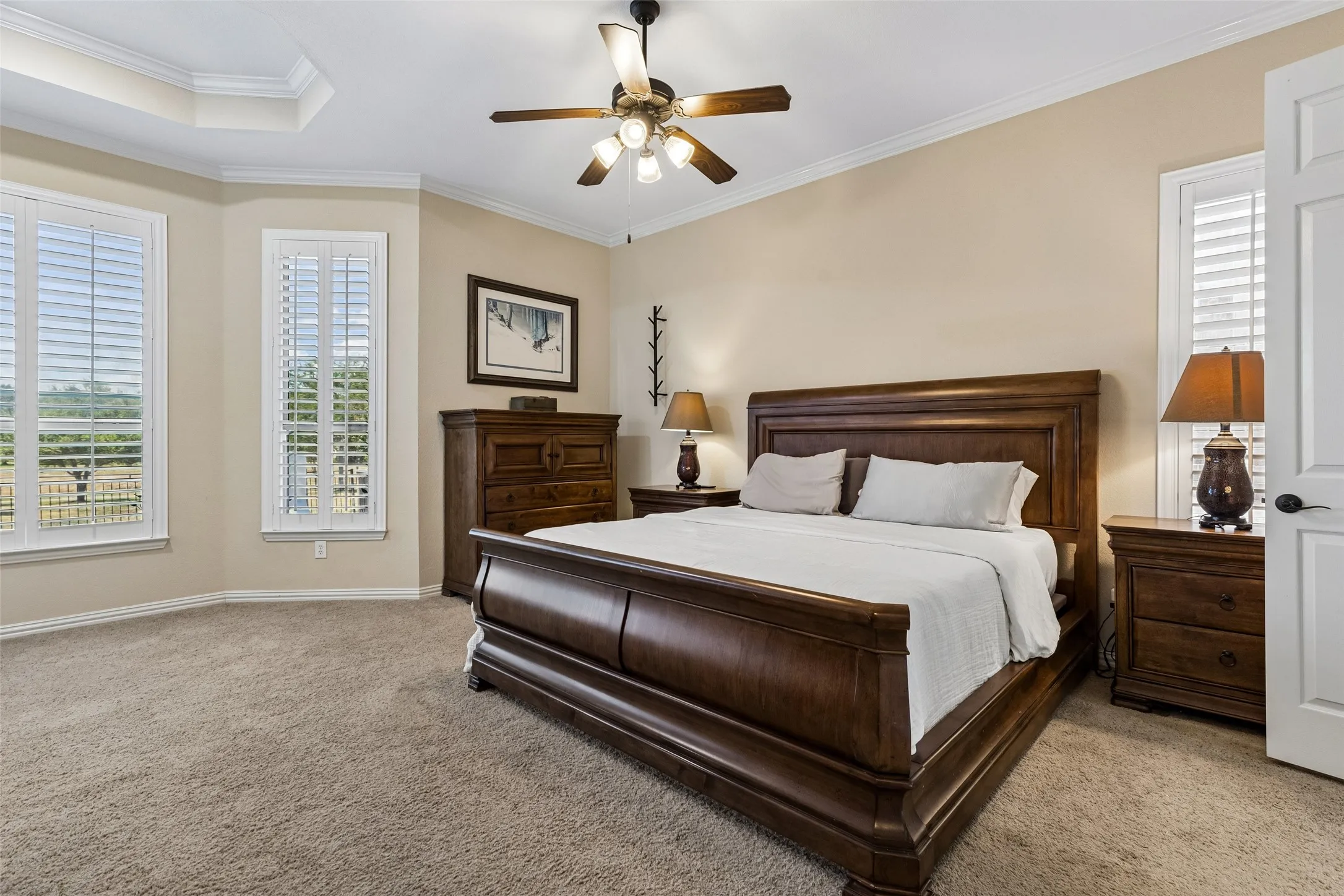Recessed tray ceiling with crown molding, three large windows with plantation shutters, ceiling fan with integrated lighting, and neutral wall tones