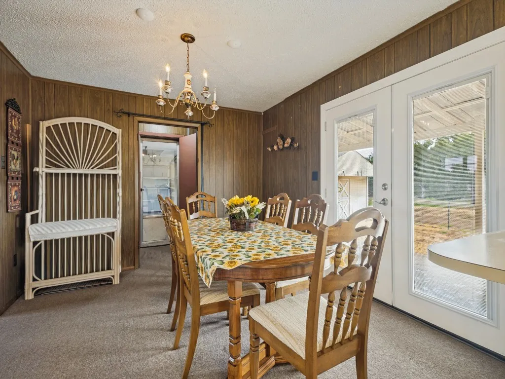The dining area features wood-panel walls, carpet flooring, a vintage chandelier, and double French doors.  The door in the background opens up to the utility room.